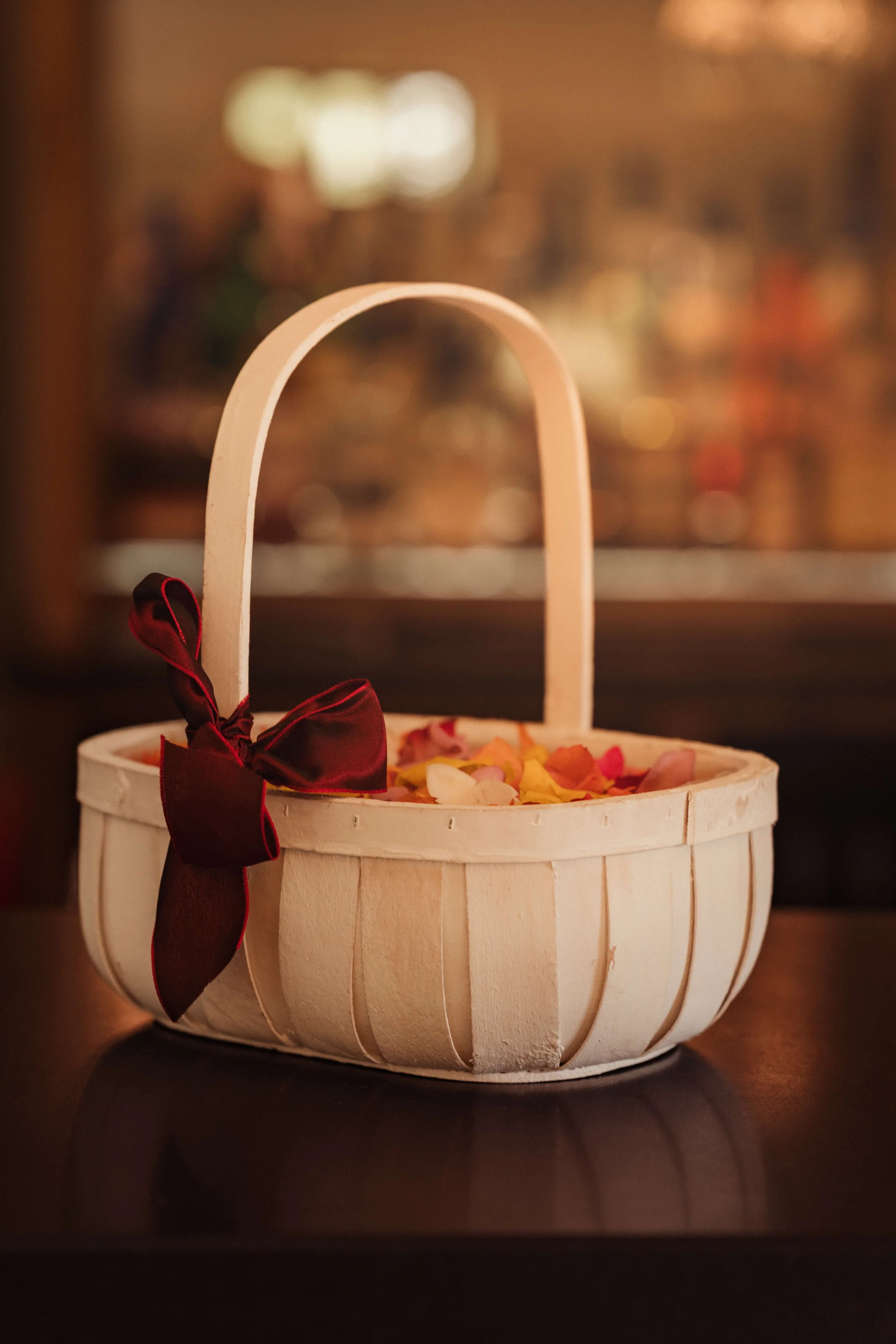 A white wicker basket with a handle and a dark red bow, filled with colorful rose petals, placed on a dark surface with a blurred warm background.