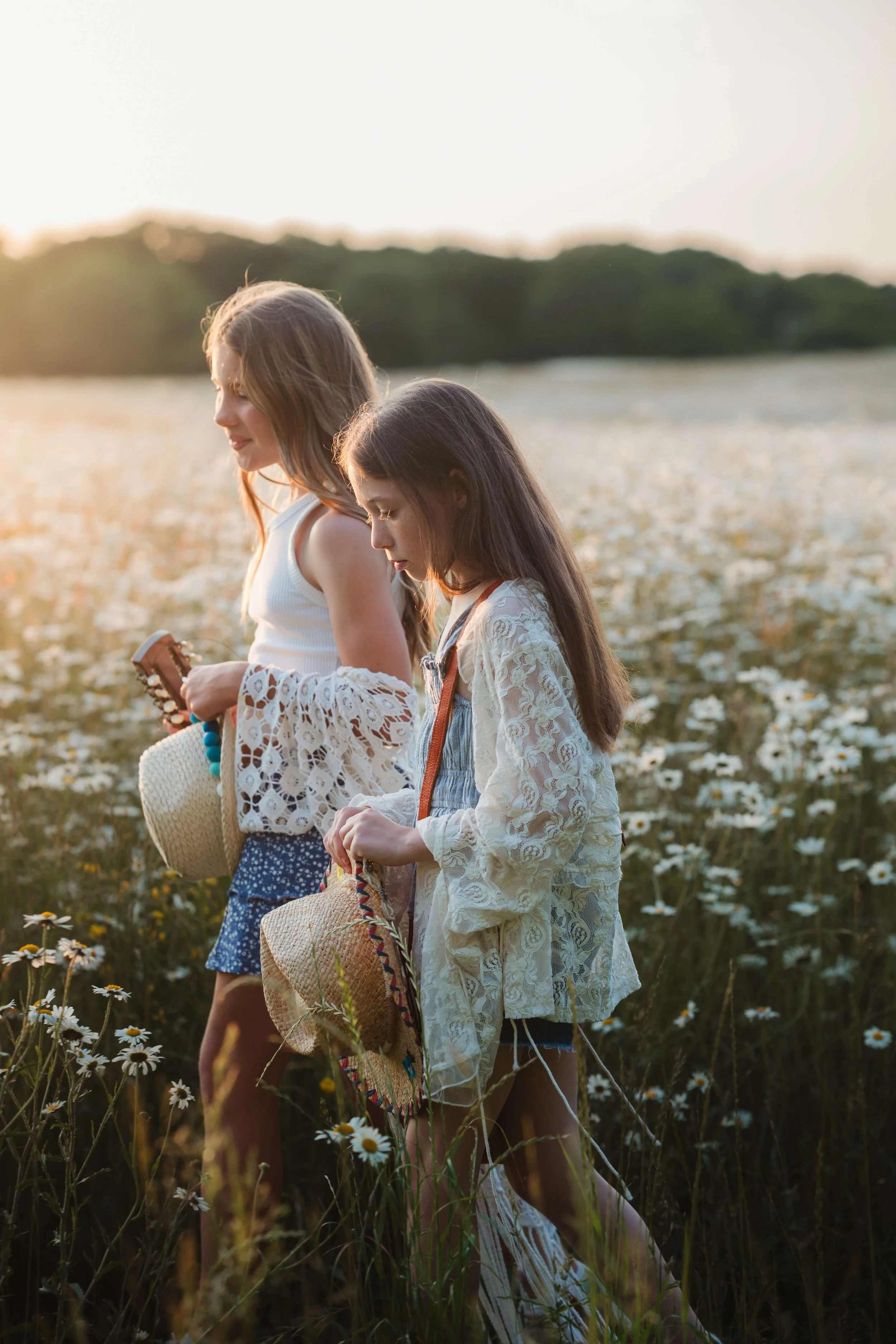 Two girls walking through a field of white daisies at sunset, holding straw hats.