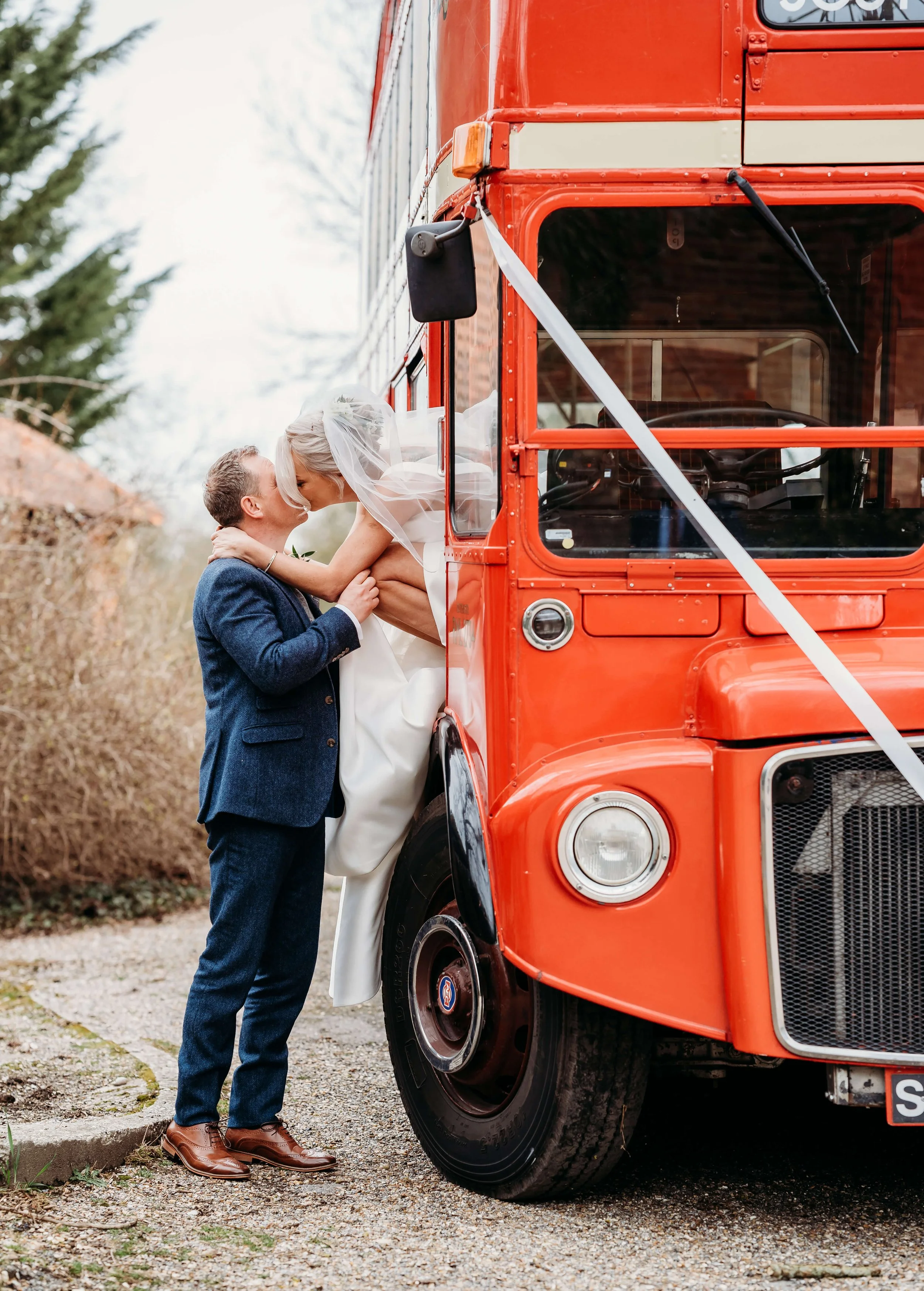A bride and groom kiss in front of a red double-decker bus, with the bride standing inside the bus leaning out to kiss the groom.