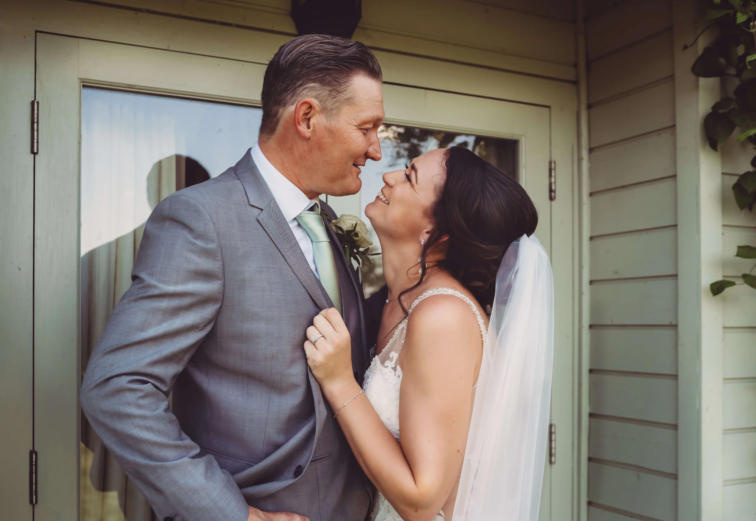 A bride and groom sharing a romantic moment outside a house, smiling and looking into each other's eyes.