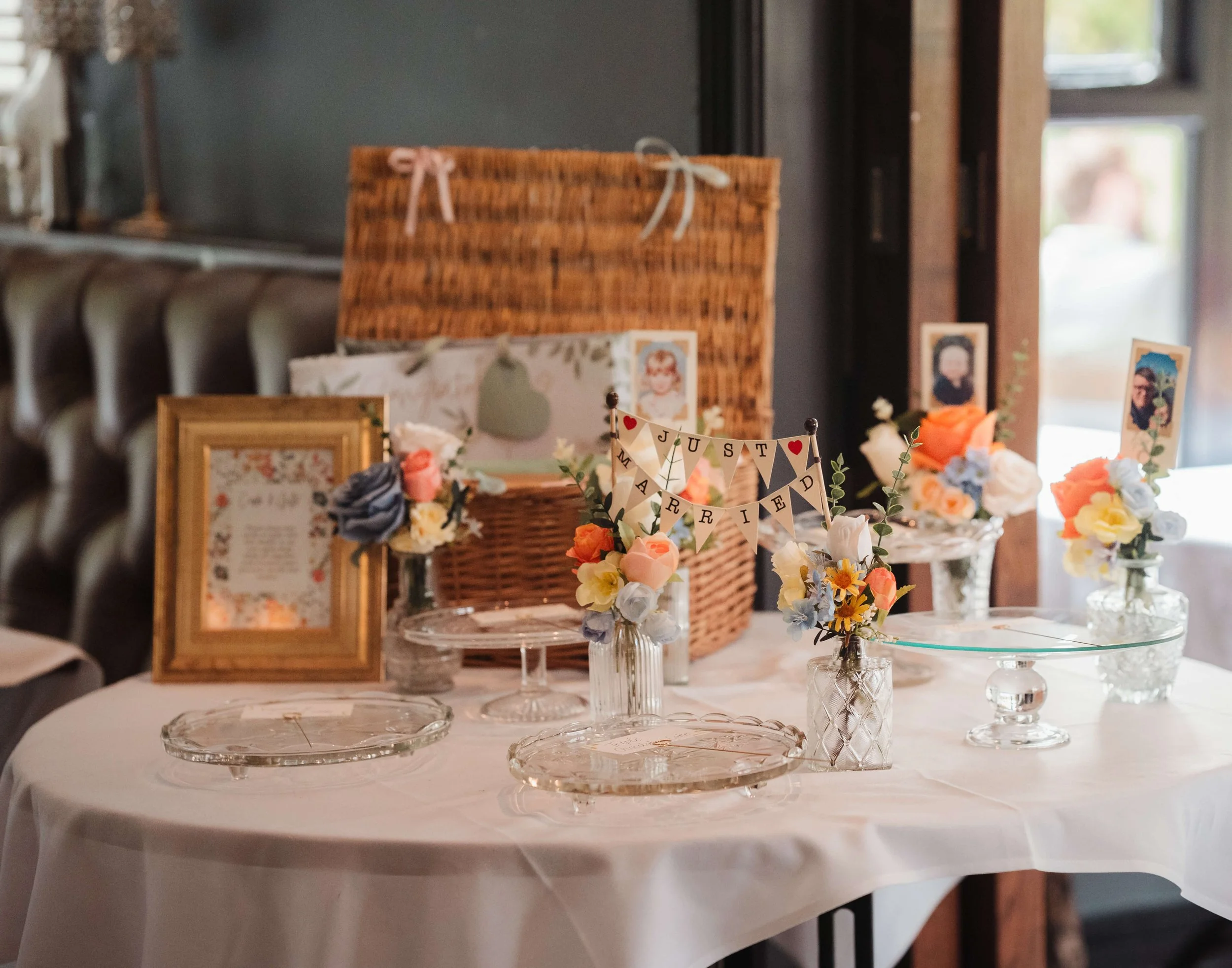 Decorative wedding table with floral arrangements, framed photo, a sign that reads "Just Married," and a basket for cards, set in a cozy indoor space.
