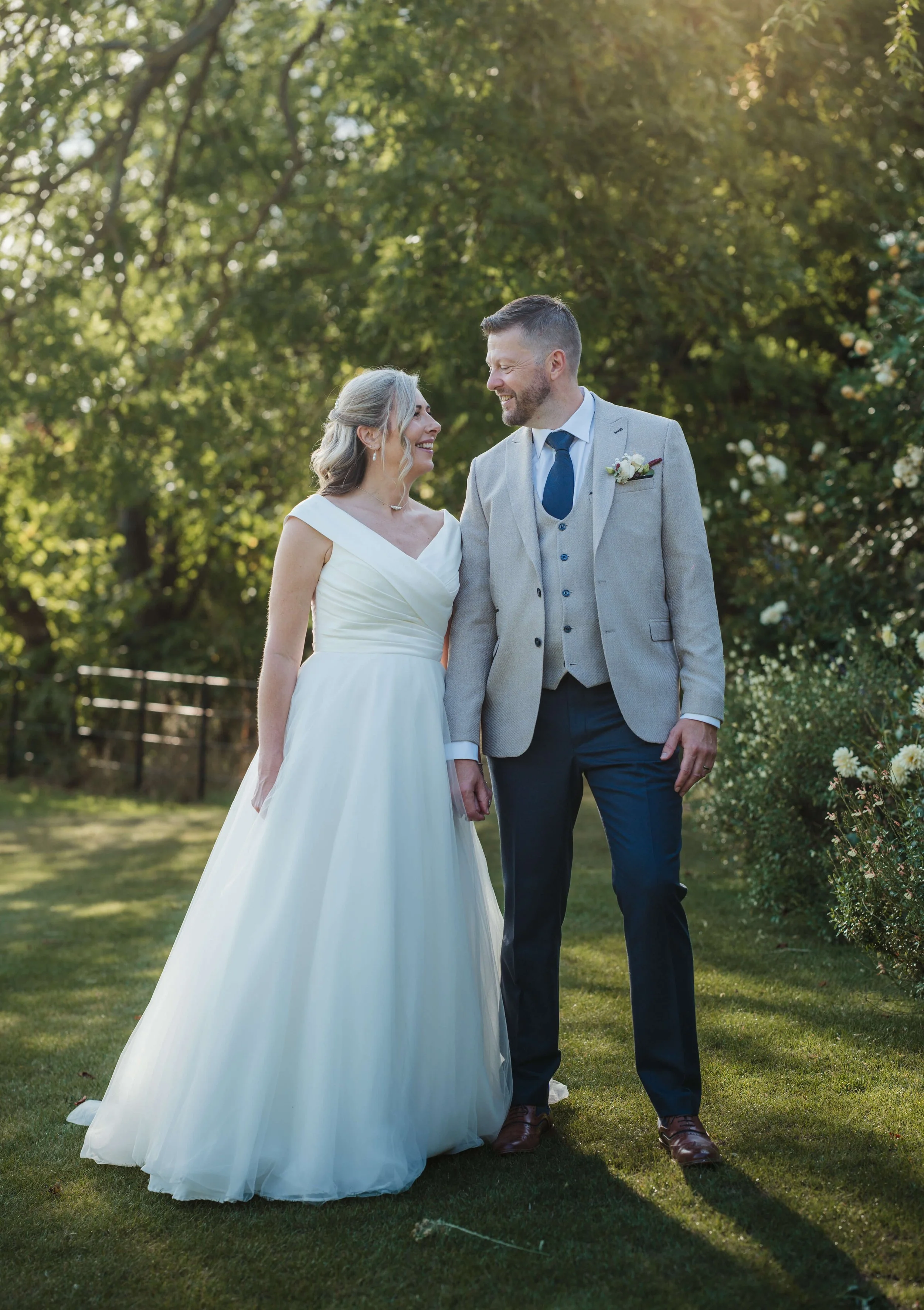 A bride and groom holding hands and smiling at each other outdoors on a sunny day, with trees and flowers in the background.