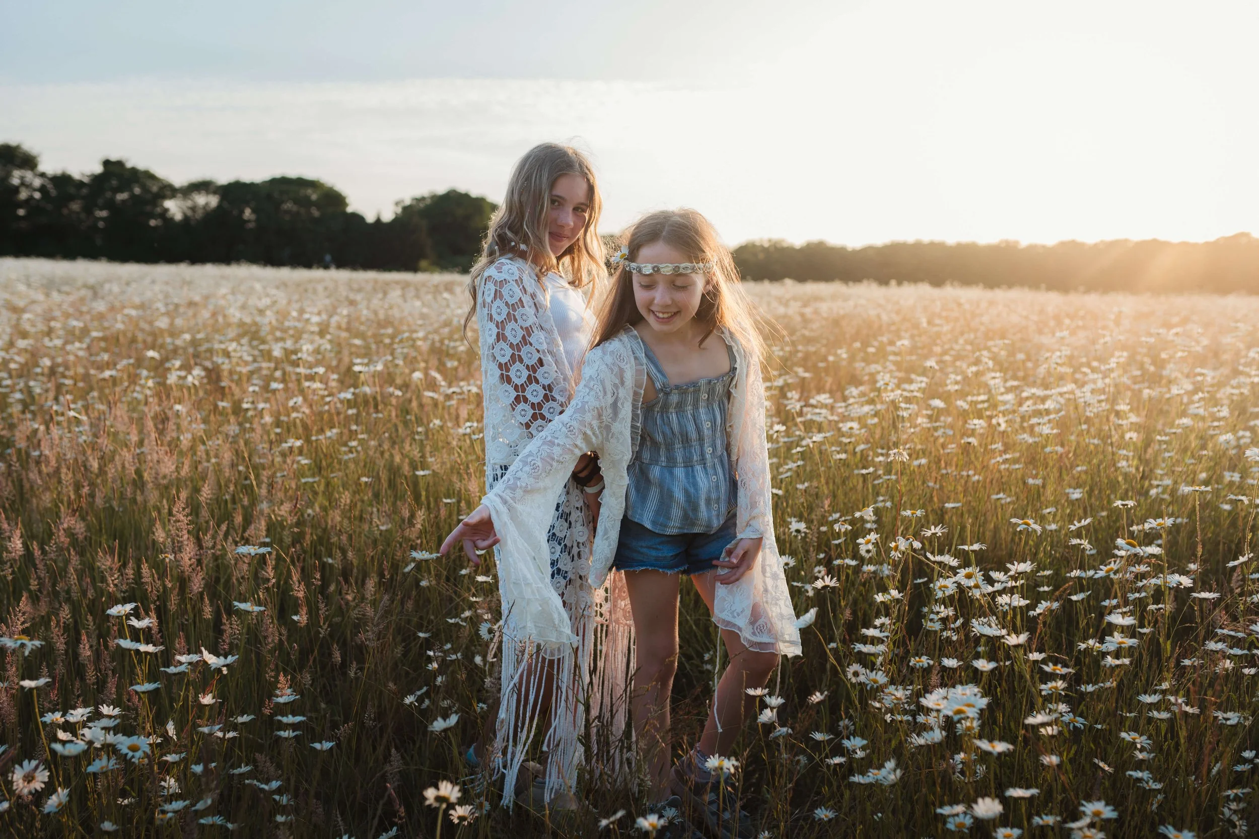 Two girls wearing bohemian-style clothing standing in a field of daisies during sunset.