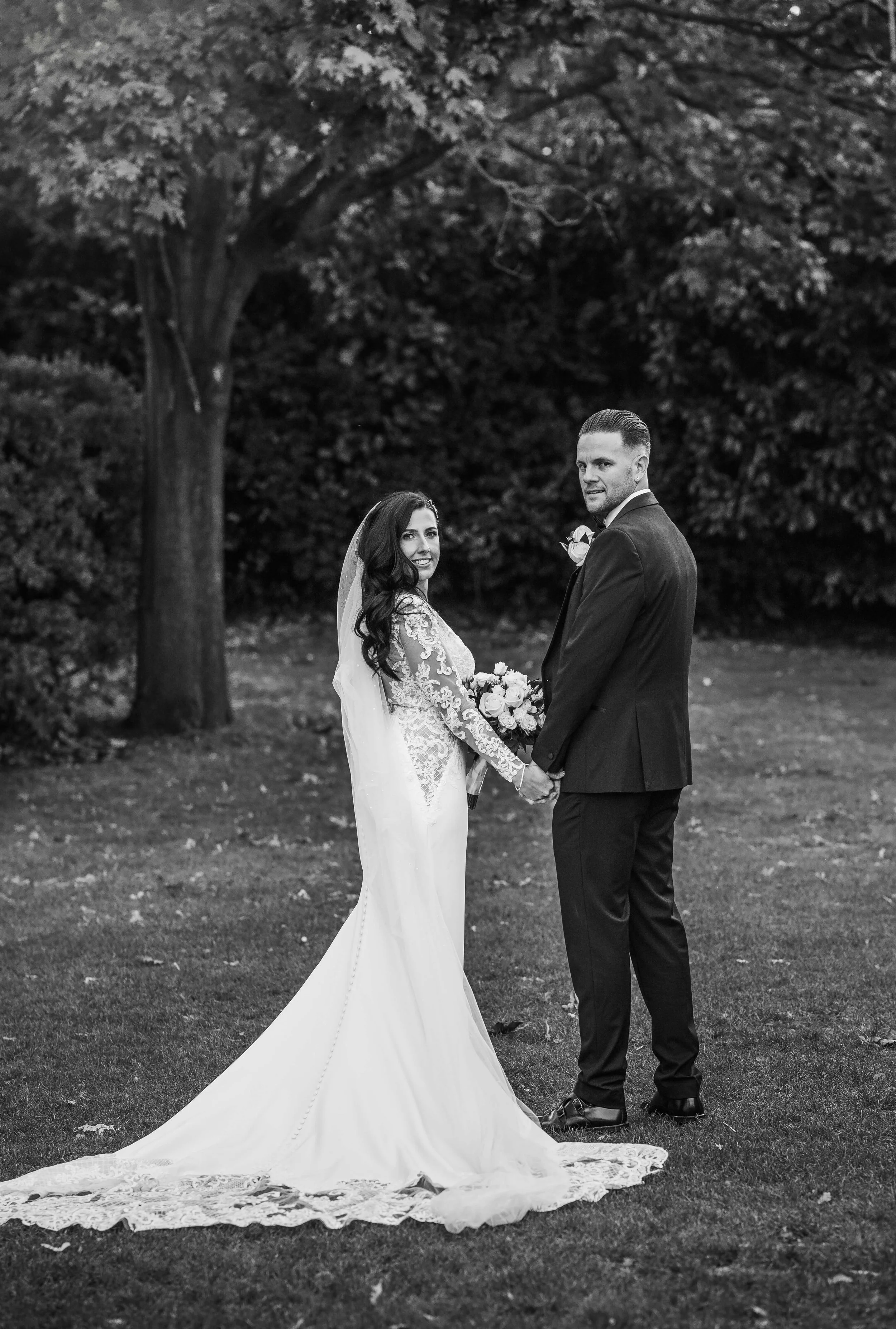 A bride and groom holding hands outdoors, dressed in wedding attire, with trees in the background.