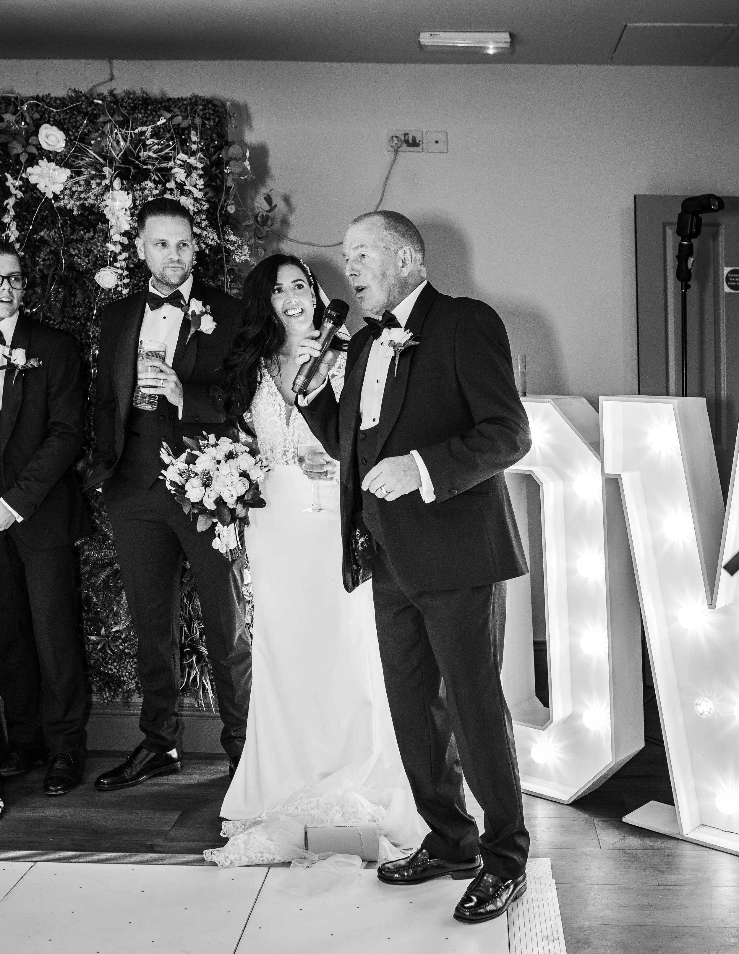 Black and white photo of a wedding celebration with a bride holding a bouquet of flowers, a groom, and a man giving a speech with a microphone. Others in tuxedos are in the background, and illuminated decorative letters are visible on the right.