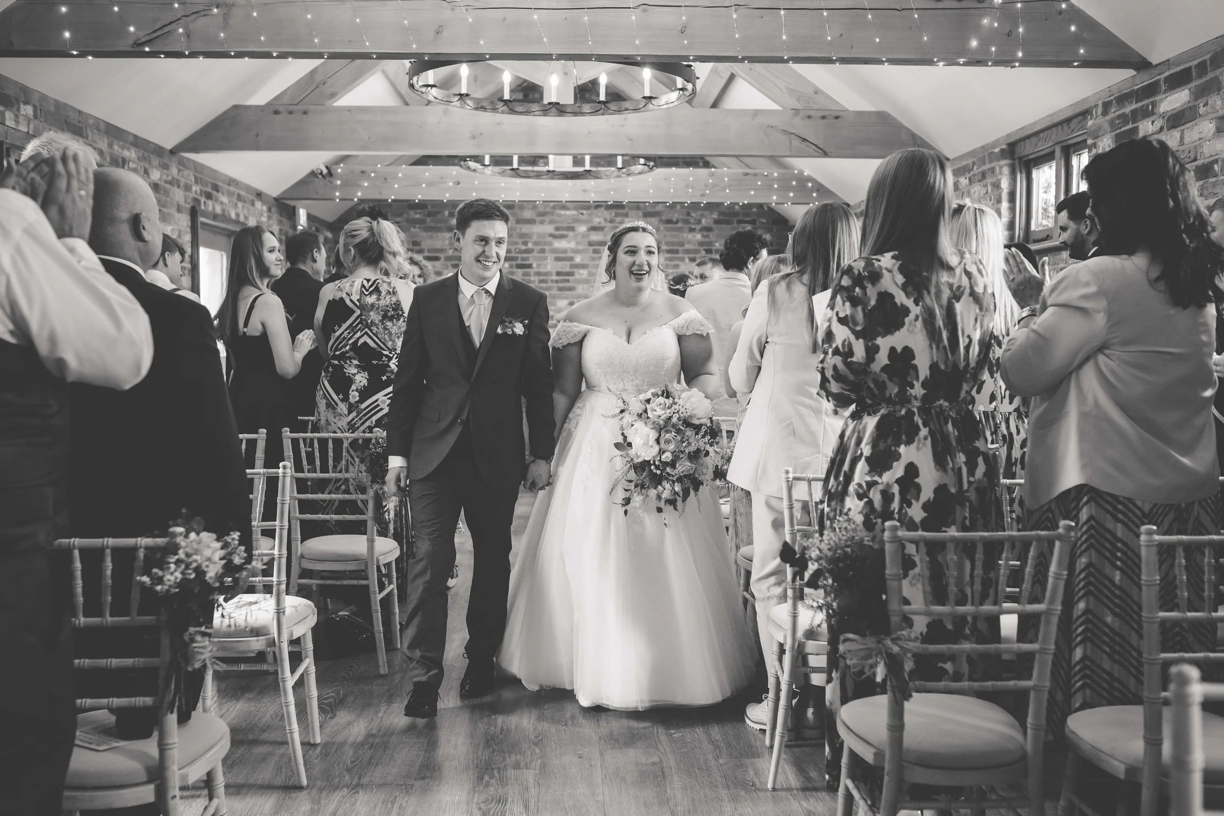 Black and white photo of a wedding reception, with a bride and groom walking down the aisle holding hands, surrounded by guests clapping and smiling.