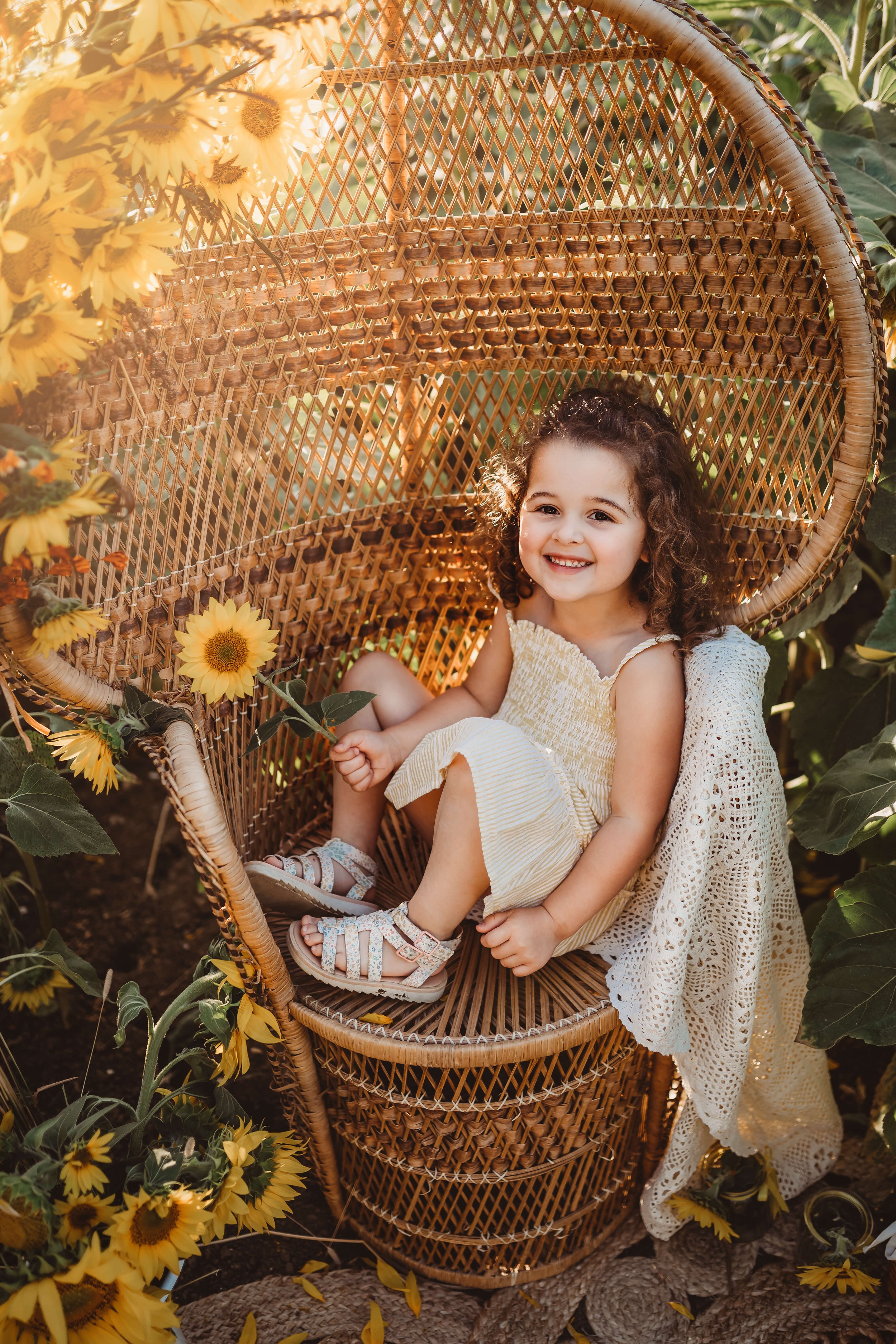 A young girl with curly hair smiling and sitting in a wicker chair outdoors surrounded by sunflowers.