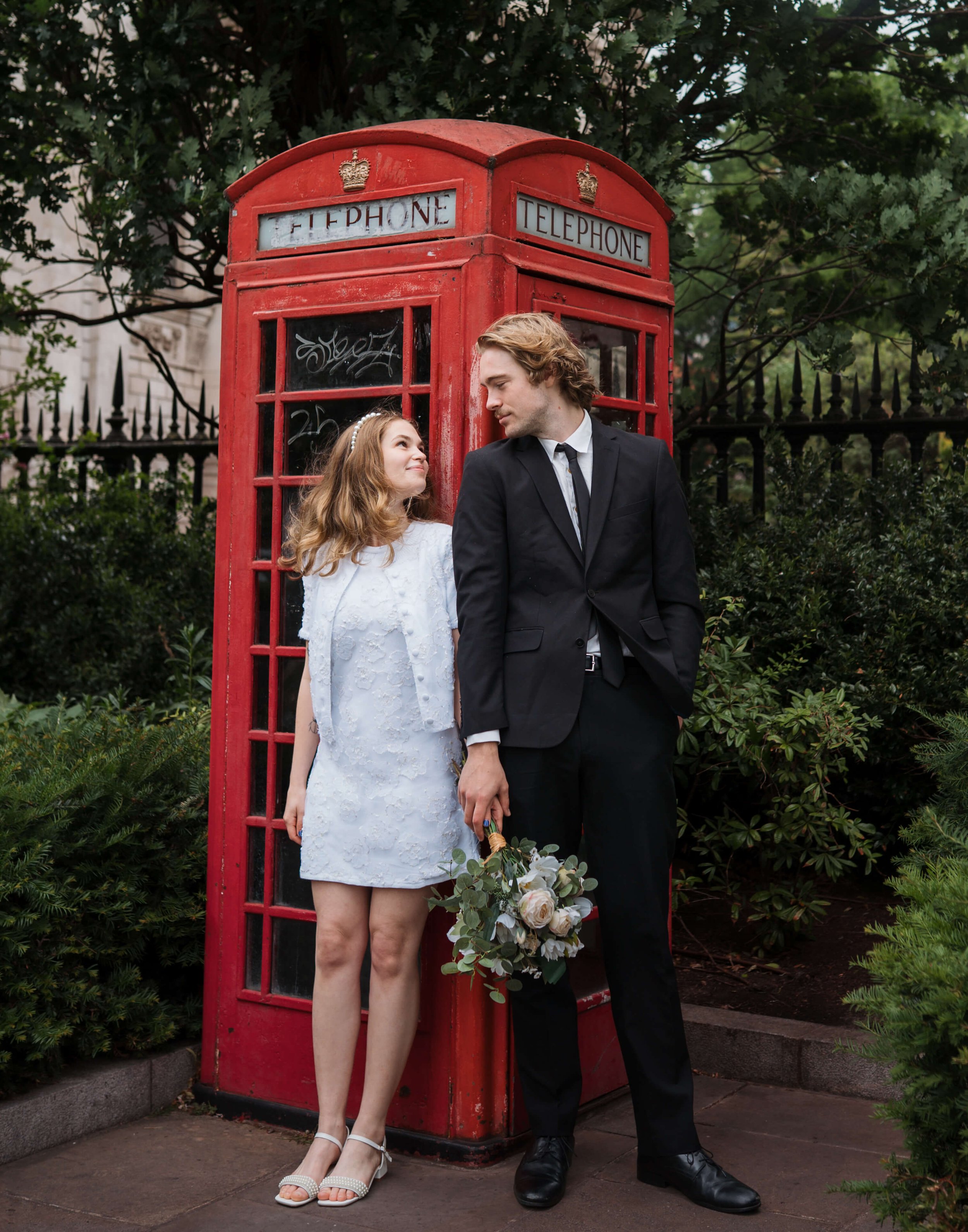 A young woman and man dressed in formal wedding attire standing in front of a red British telephone booth, with greenery in the background.