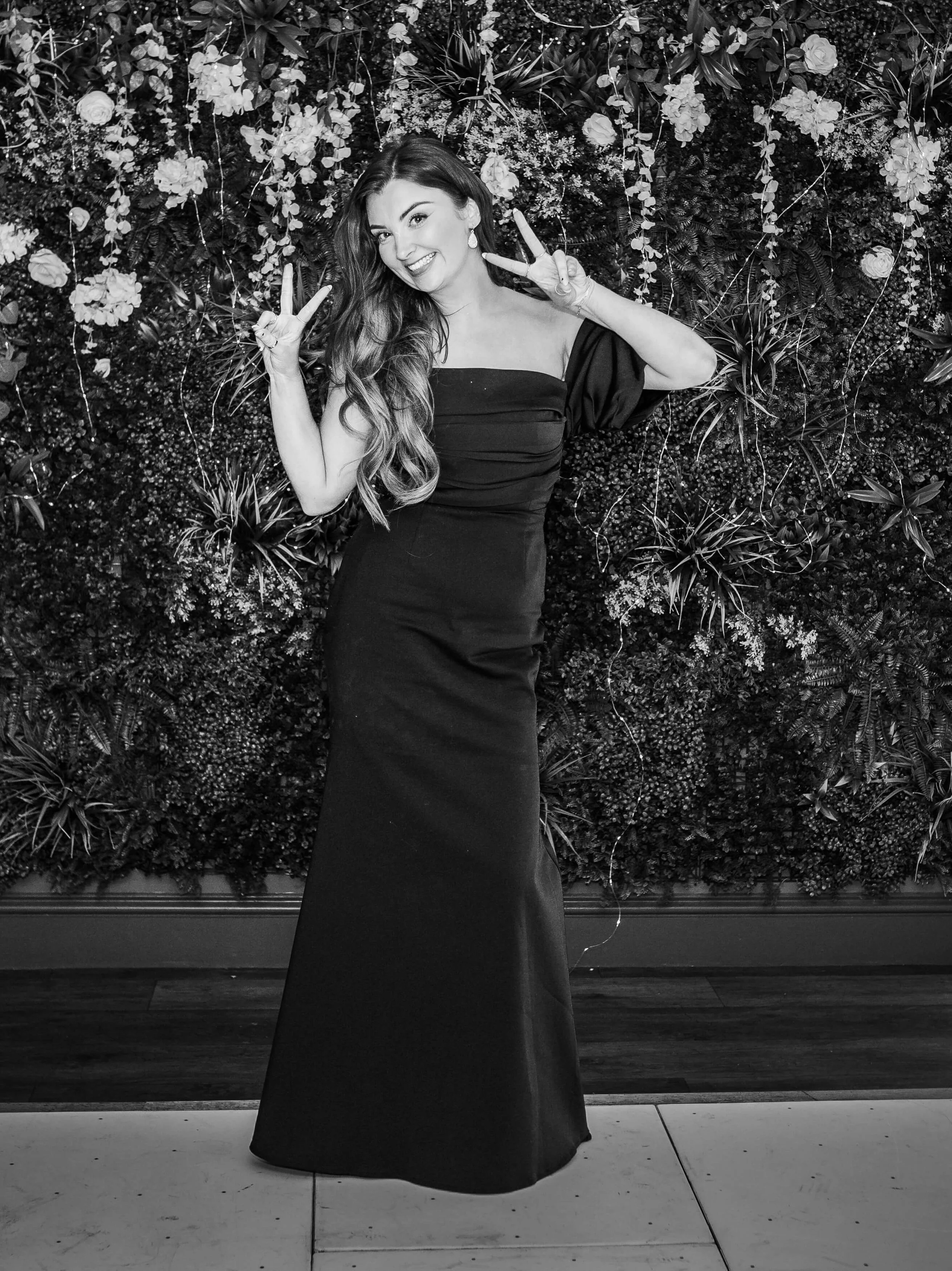 Woman in a long black dress making peace signs with both hands, standing in front of a floral wall backdrop.