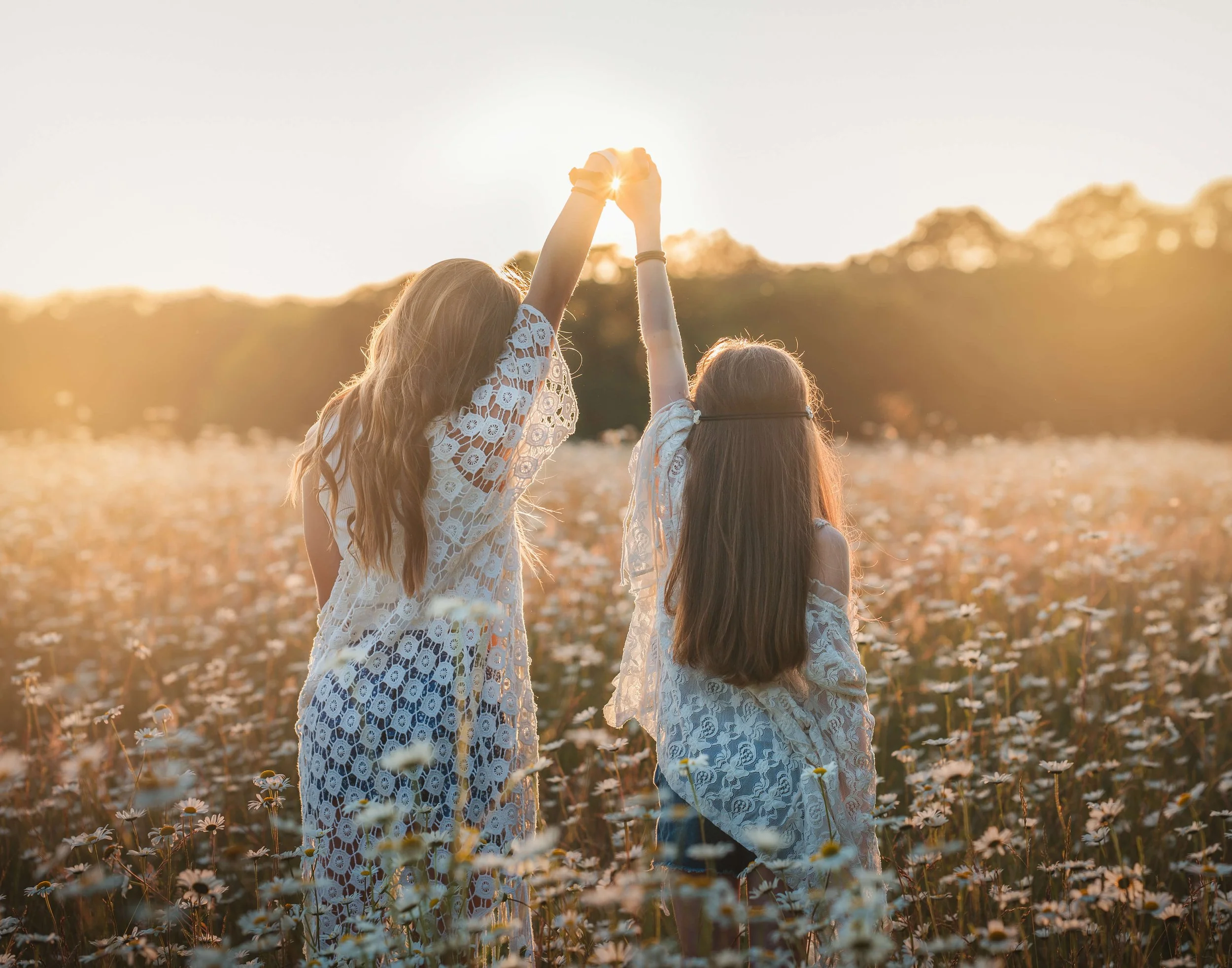 Two girls in white lace dresses holding hands and raising them together in a field of flowers during sunset.