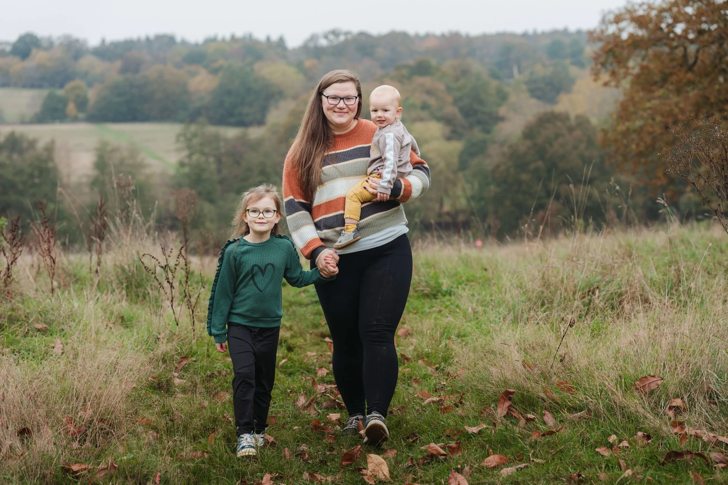 A woman walking outdoors with two children, holding their hands, in a grassy field with fall foliage in the background.