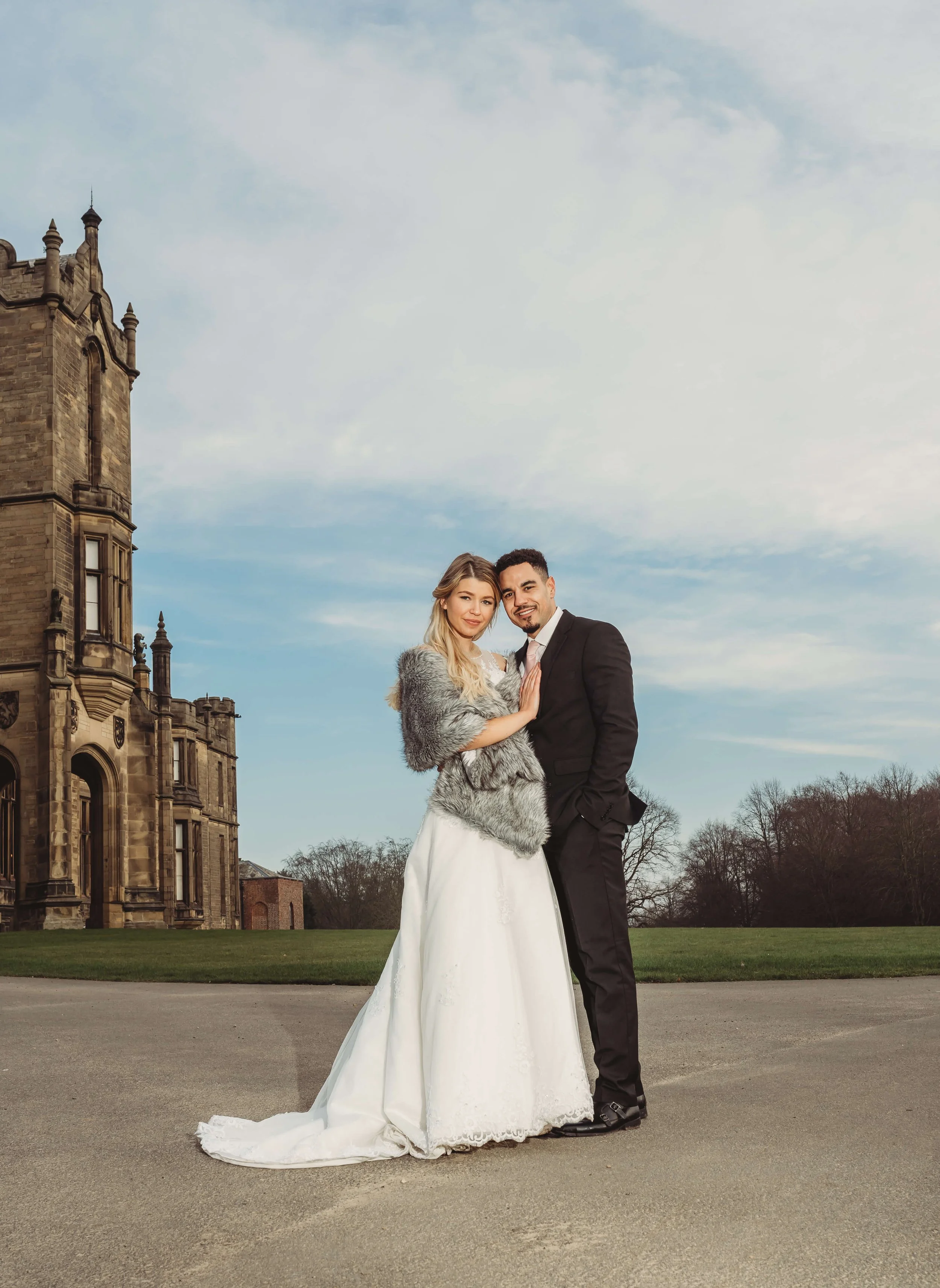A newlywed couple stands outdoors in front of a historic castle-like building, with a cloudy sky above and trees in the background. The bride wears a white wedding gown and a grey fur shawl, while the groom is dressed in a black suit and tie.