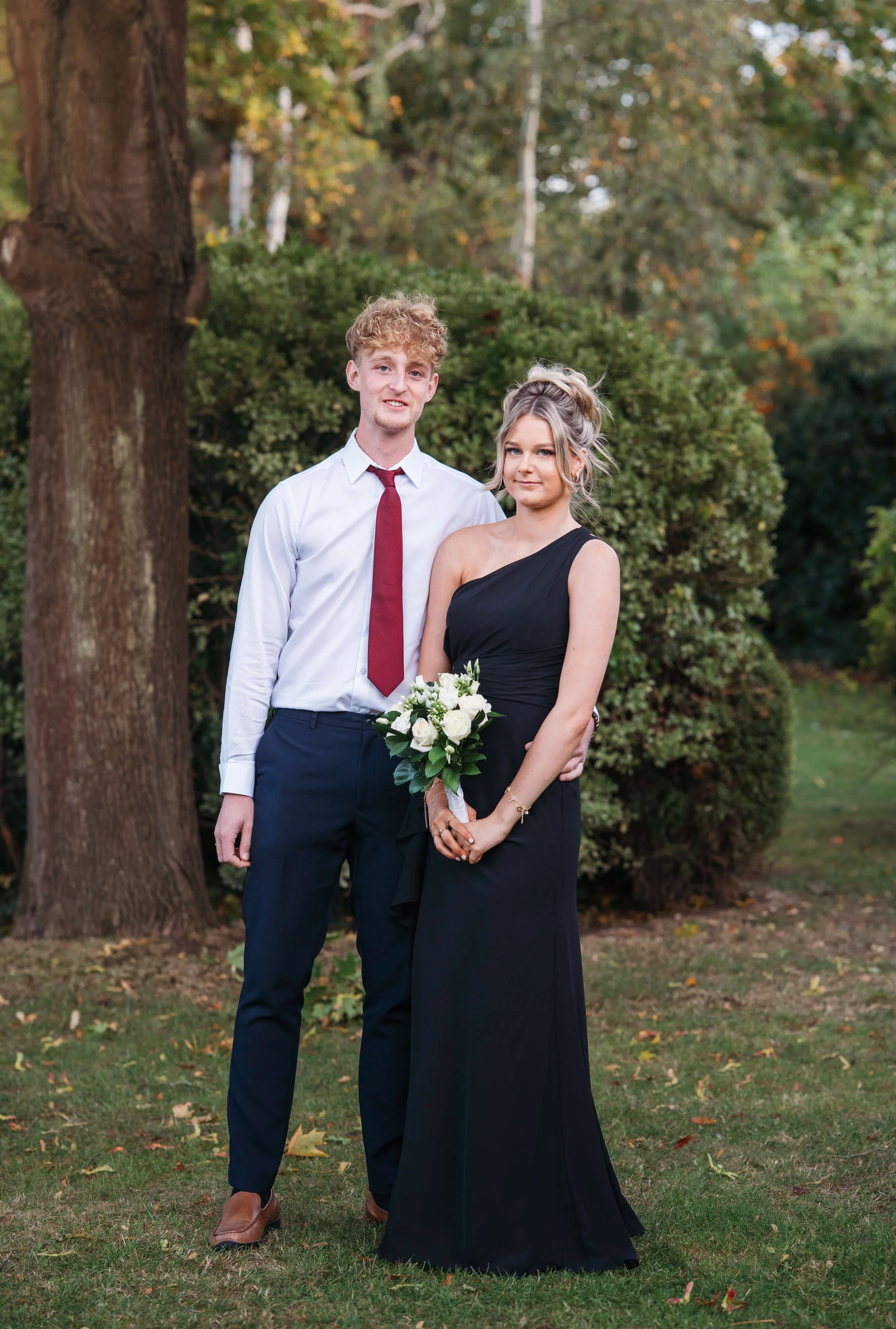 A young man in a white shirt, navy pants, and red tie stands next to a young woman in a black dress holding a bouquet of white roses, outdoors in front of trees and shrubs.