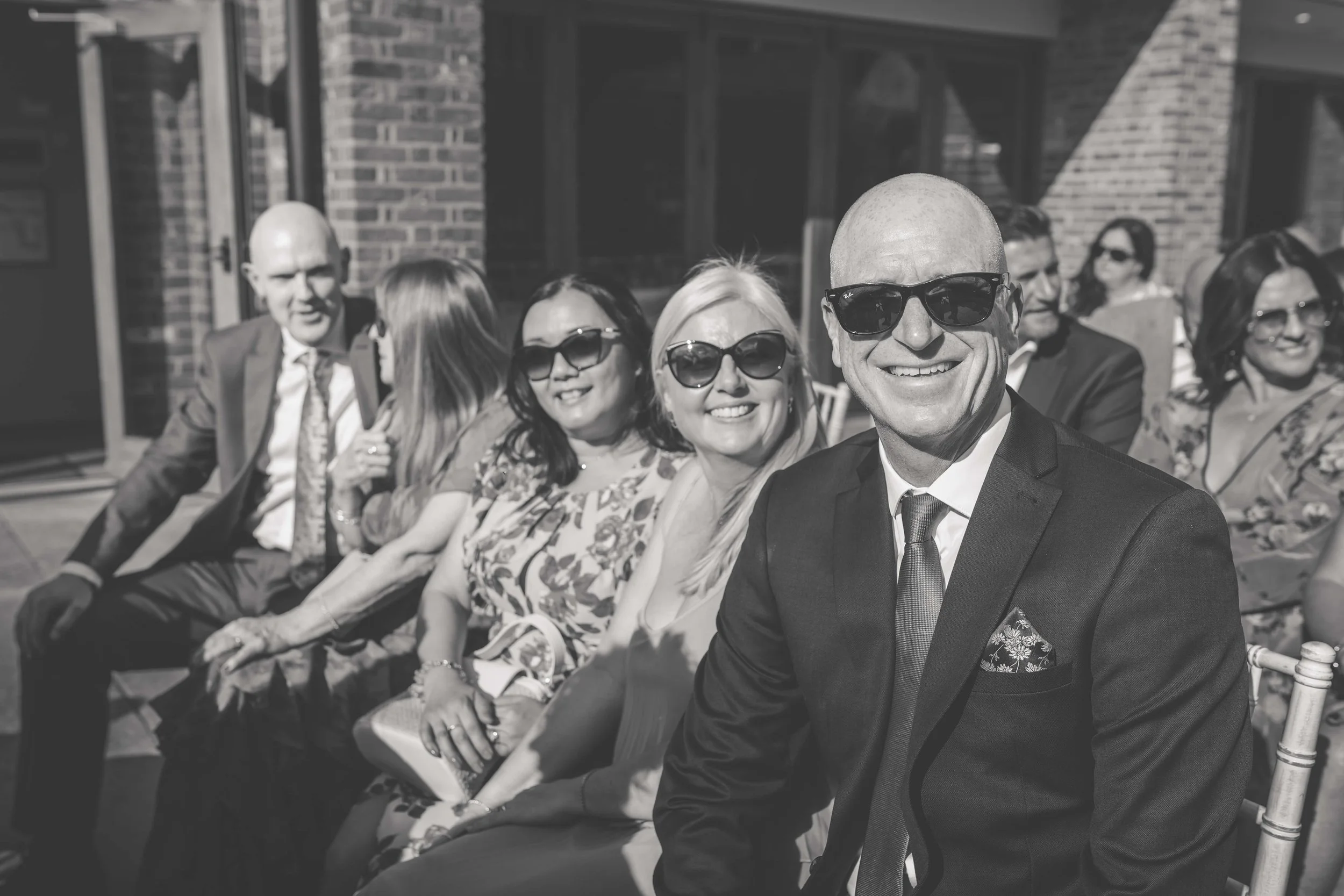 A group of formally dressed adults sitting outdoors on a sunny day, smiling and wearing sunglasses.