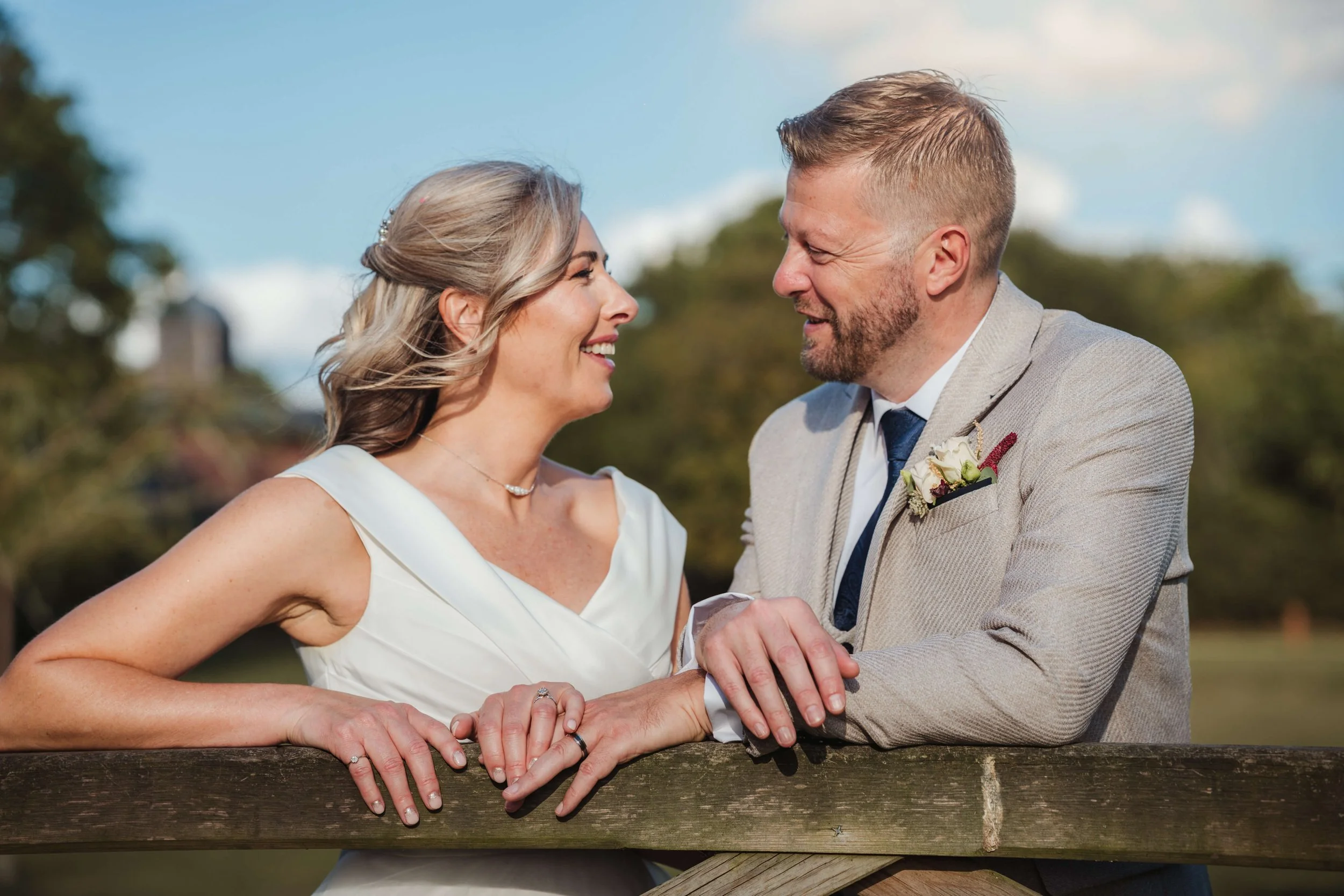A couple on their wedding day smiling at each other outdoors, holding hands on a wooden bridge, with trees and a cloudy sky in the background.
