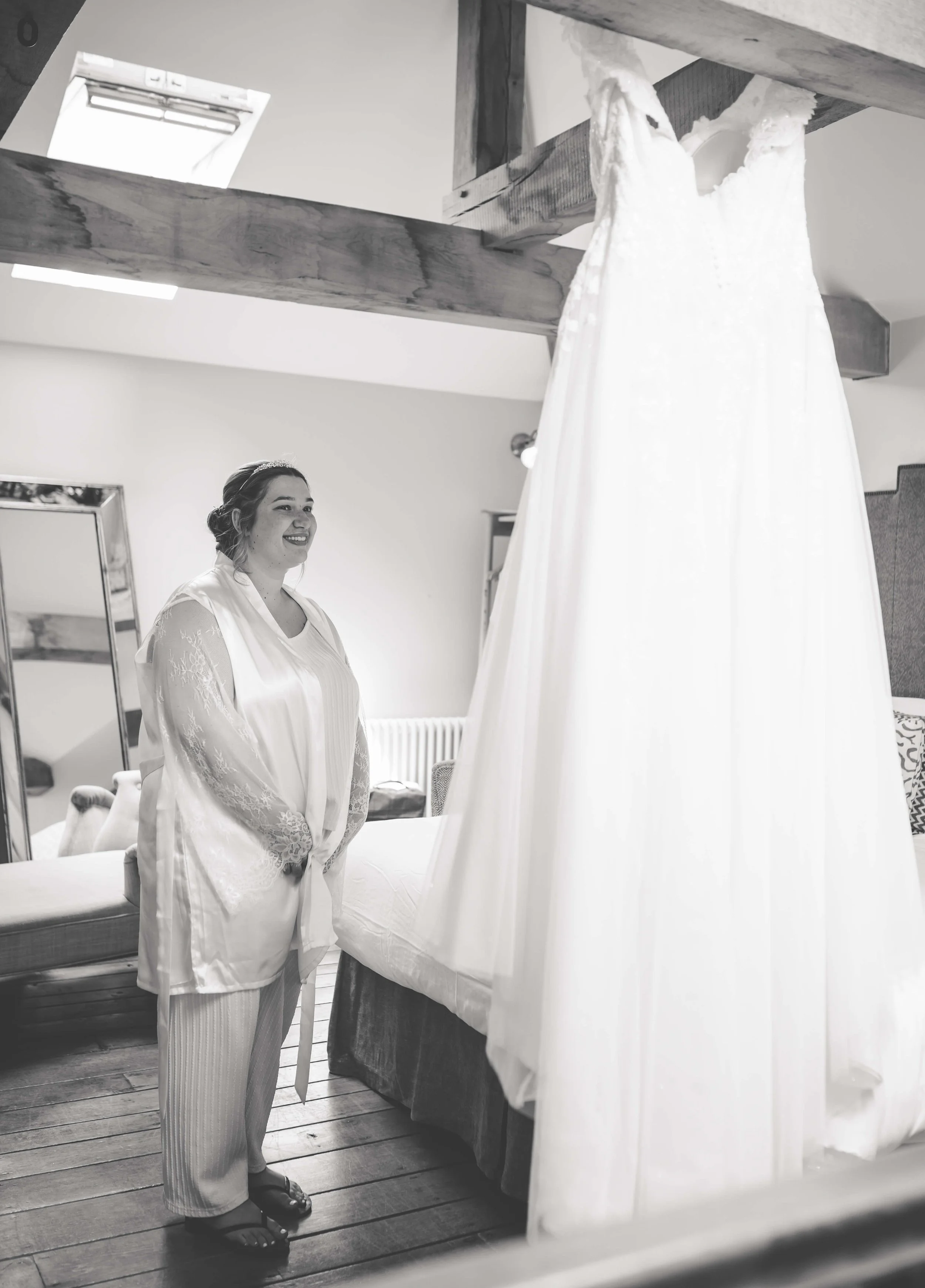 A woman in pajamas standing and smiling in a bedroom, looking at a white dress hanging over a bed