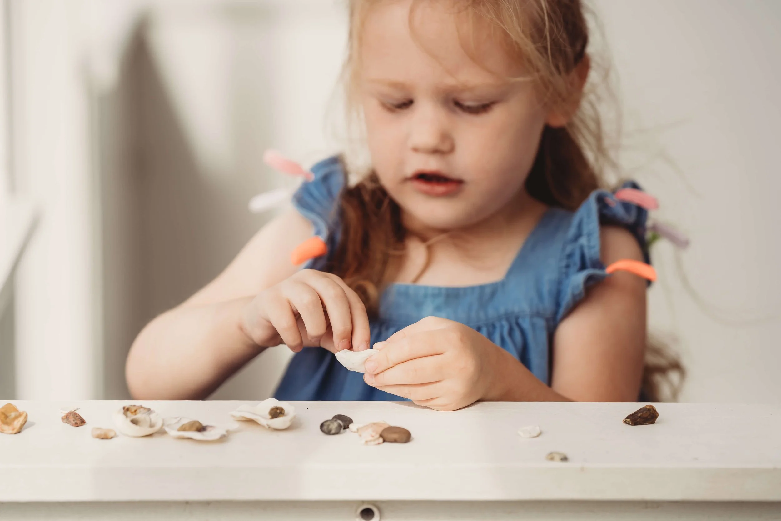 A young girl with curly hair, wearing a blue dress with colorful fringe on the shoulders, sitting at a table and examining seashells.