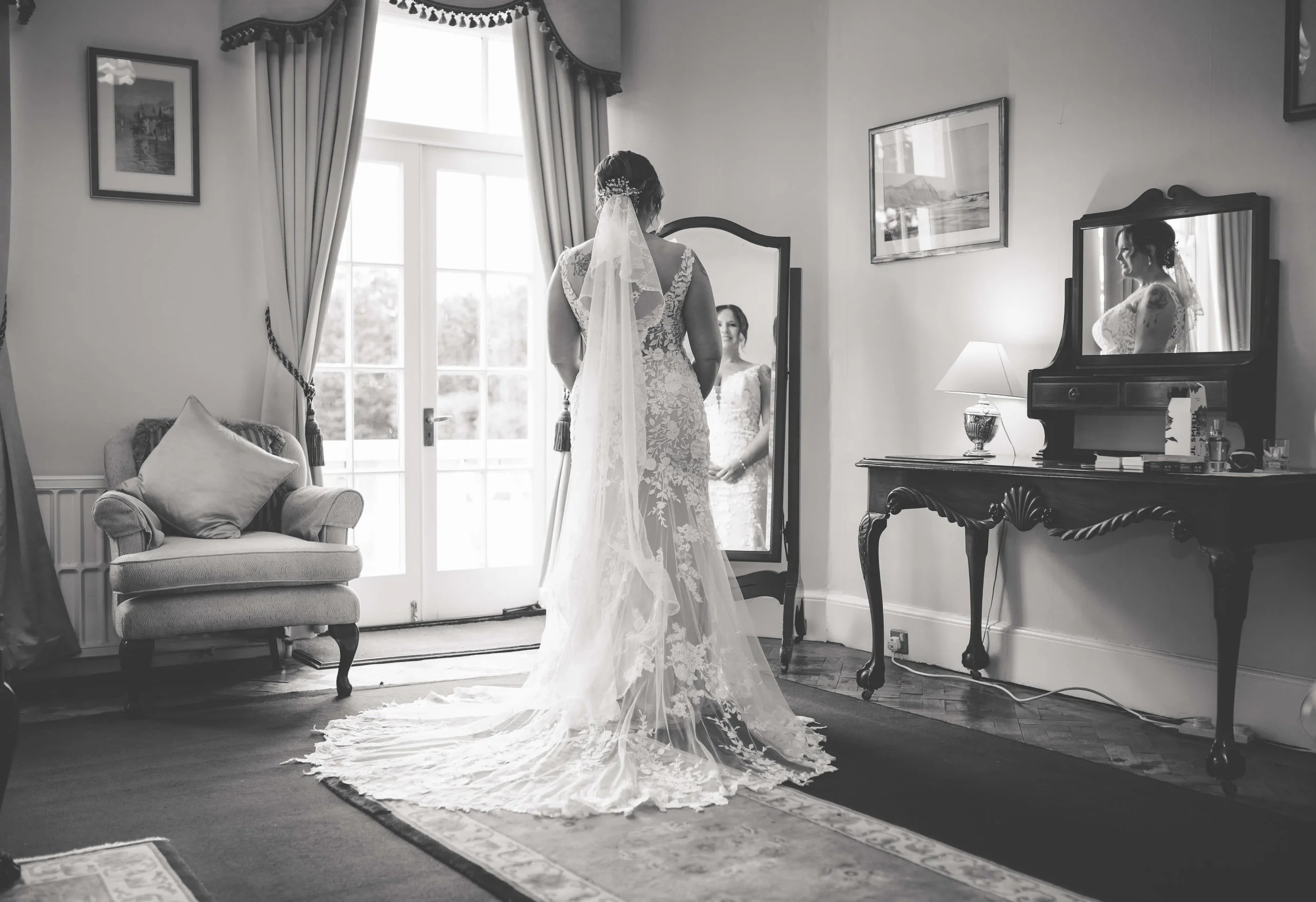 A bride in a wedding dress looking at her reflection in a full-length mirror in a well-lit, elegant room with curtains, paintings, and antique furniture.