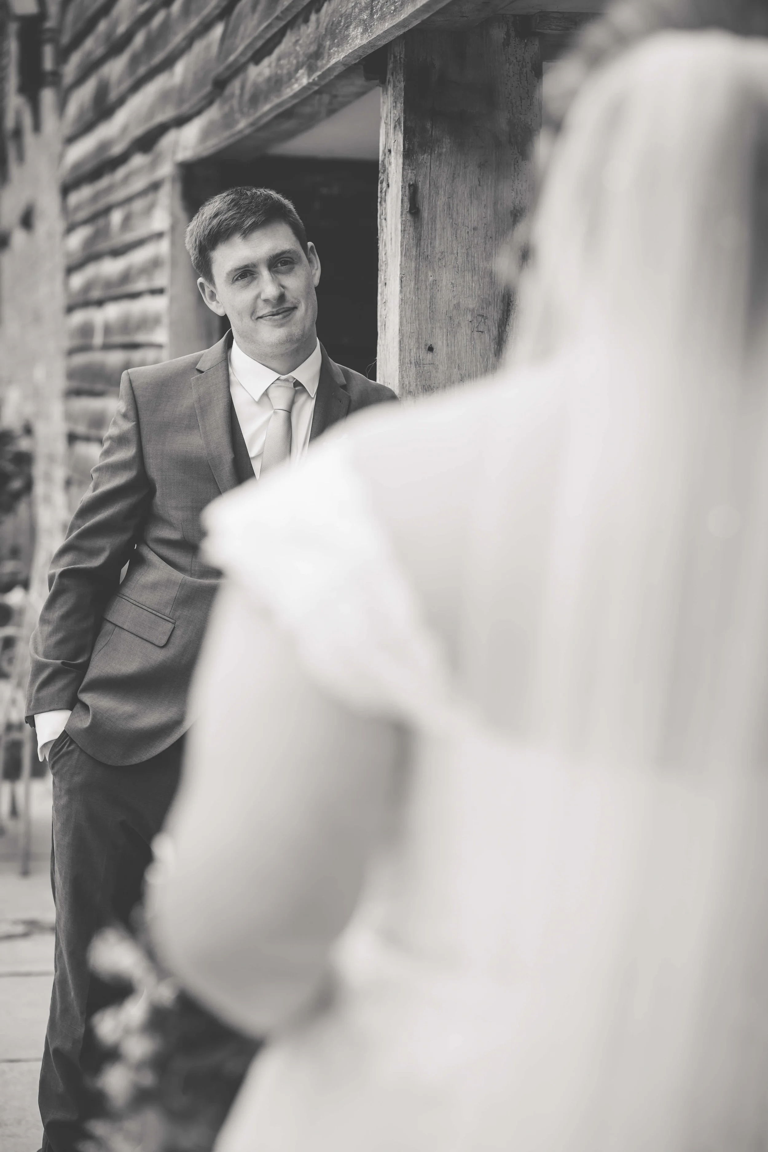 A man in a suit looking at a woman, standing outside near a wooden structure.
