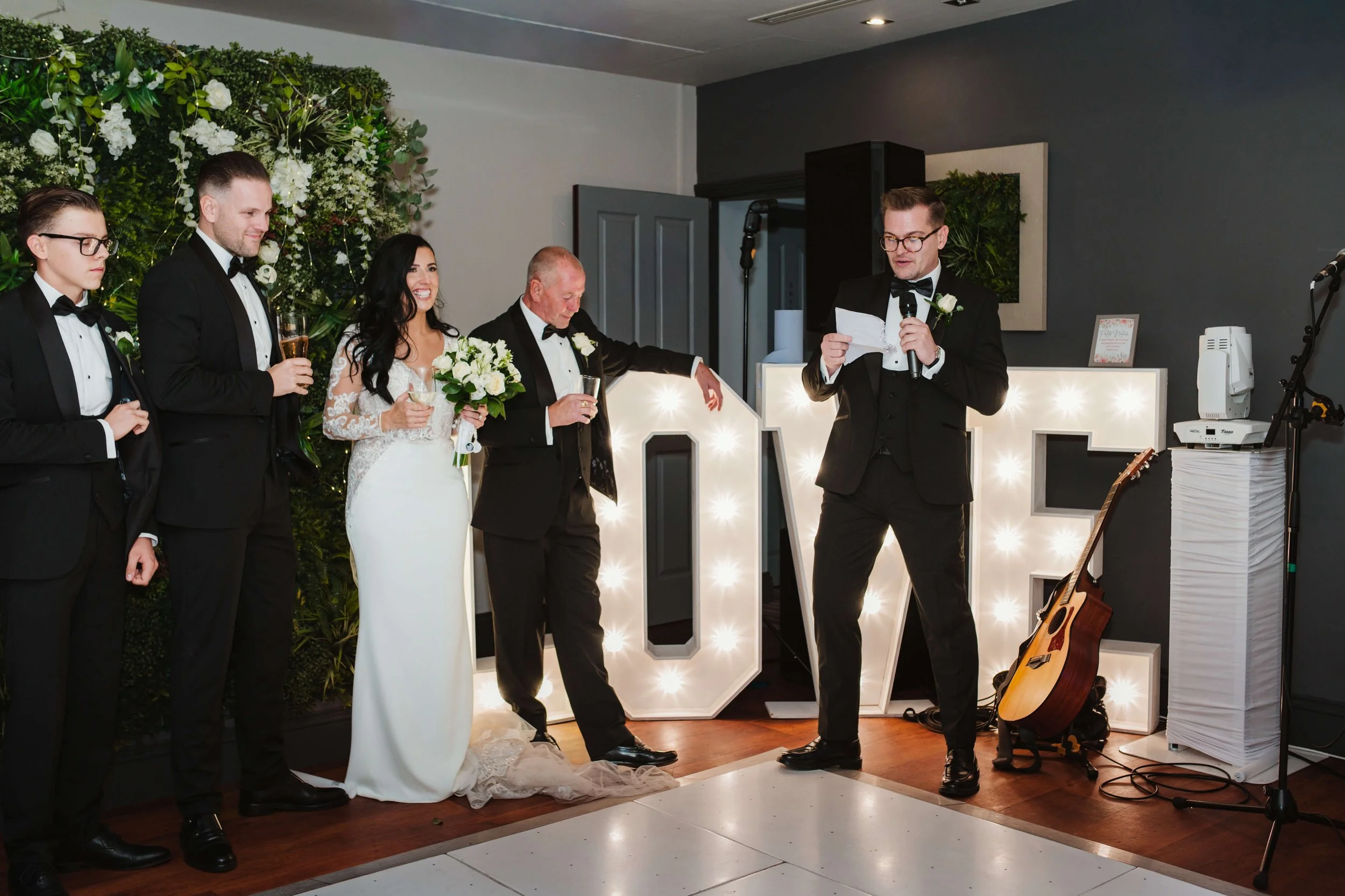 A bride and groom stand with members of the wedding party, all dressed in formal attire, during a wedding celebration. The groom is giving a speech or toast, holding a microphone and reading from a paper, while the bride holds a bouquet. A person to 