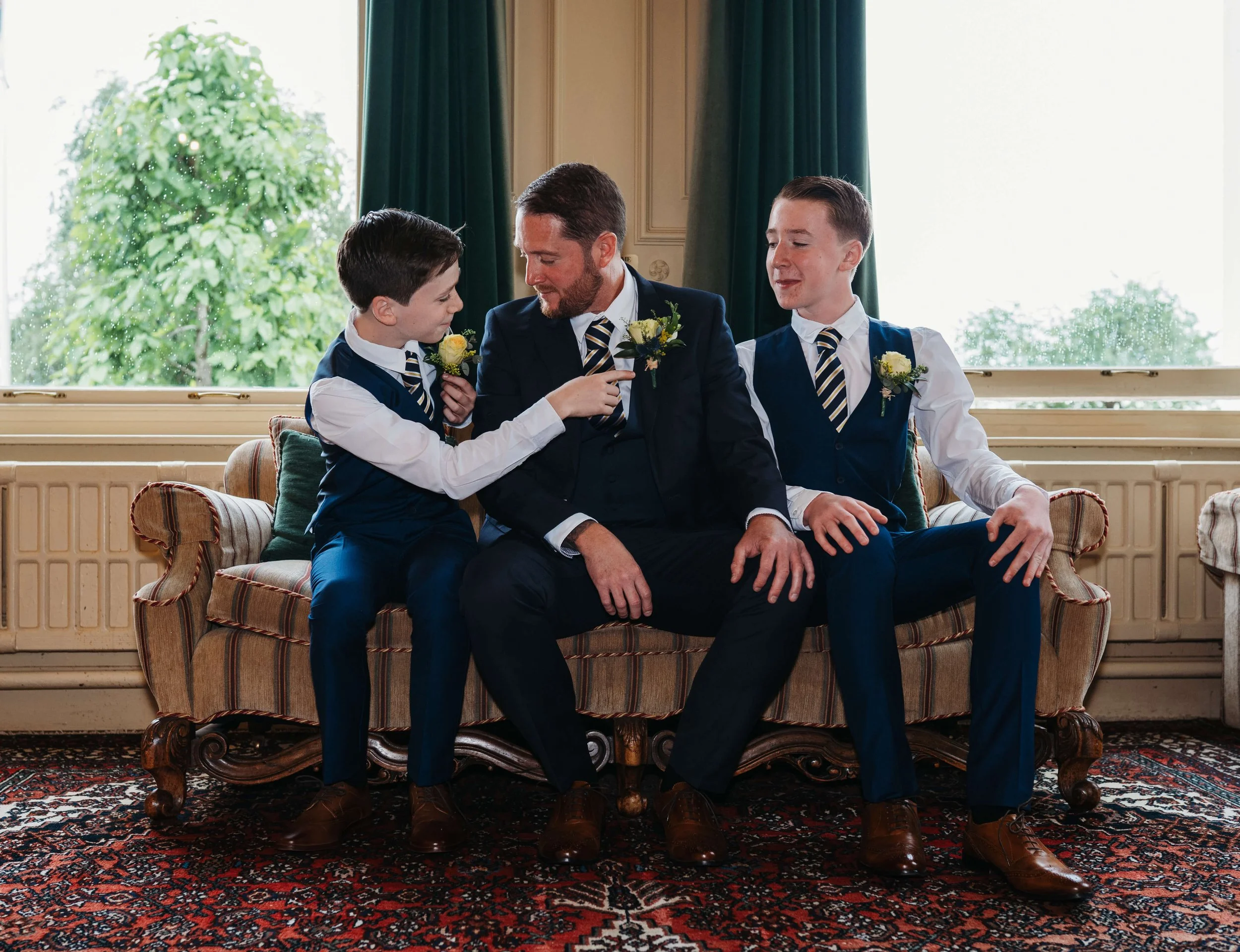 A man and two boys sitting on a sofa in a room with green curtains and a large window, dressed in formal suits with striped ties, engaging in a playful moment.