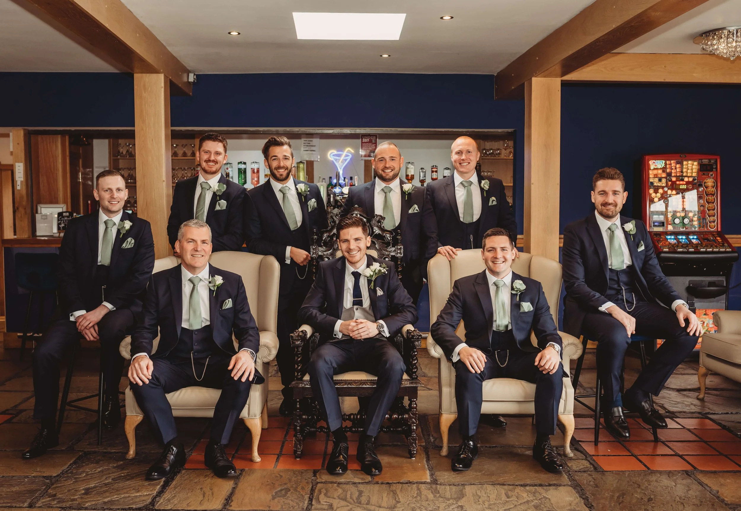 A group of nine groomsmen dressed in formal suits and ties, seated and standing in a bar or lounge area. They are smiling and  celebrating,  a wedding special event.