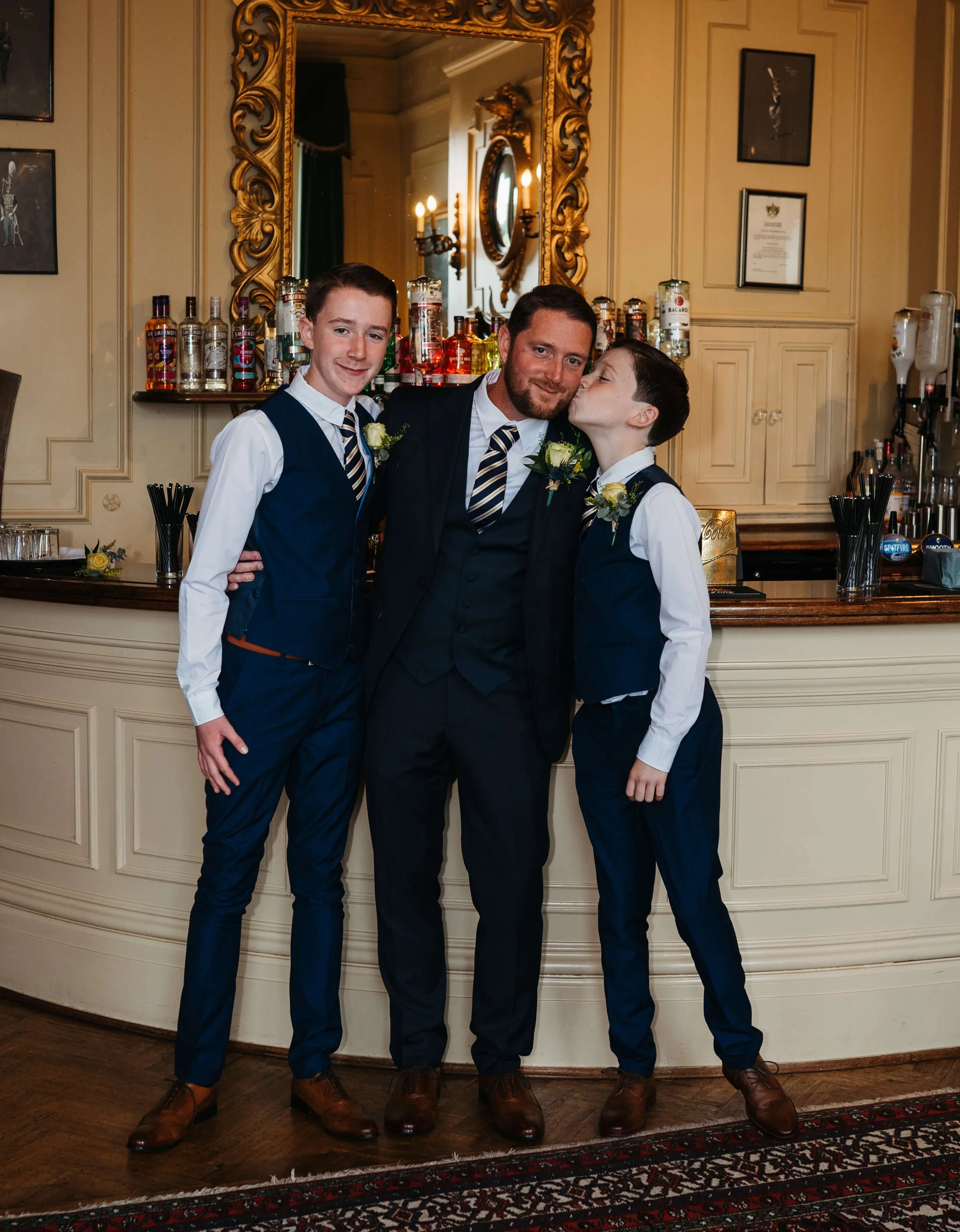 A man and two boys dressed in formal attire, standing indoors in front of a bar with bottles of alcohol, a large ornate mirror, and framed certificates on the wall. The man is kissing the boy on the right, and all three are smiling. They are wearing 