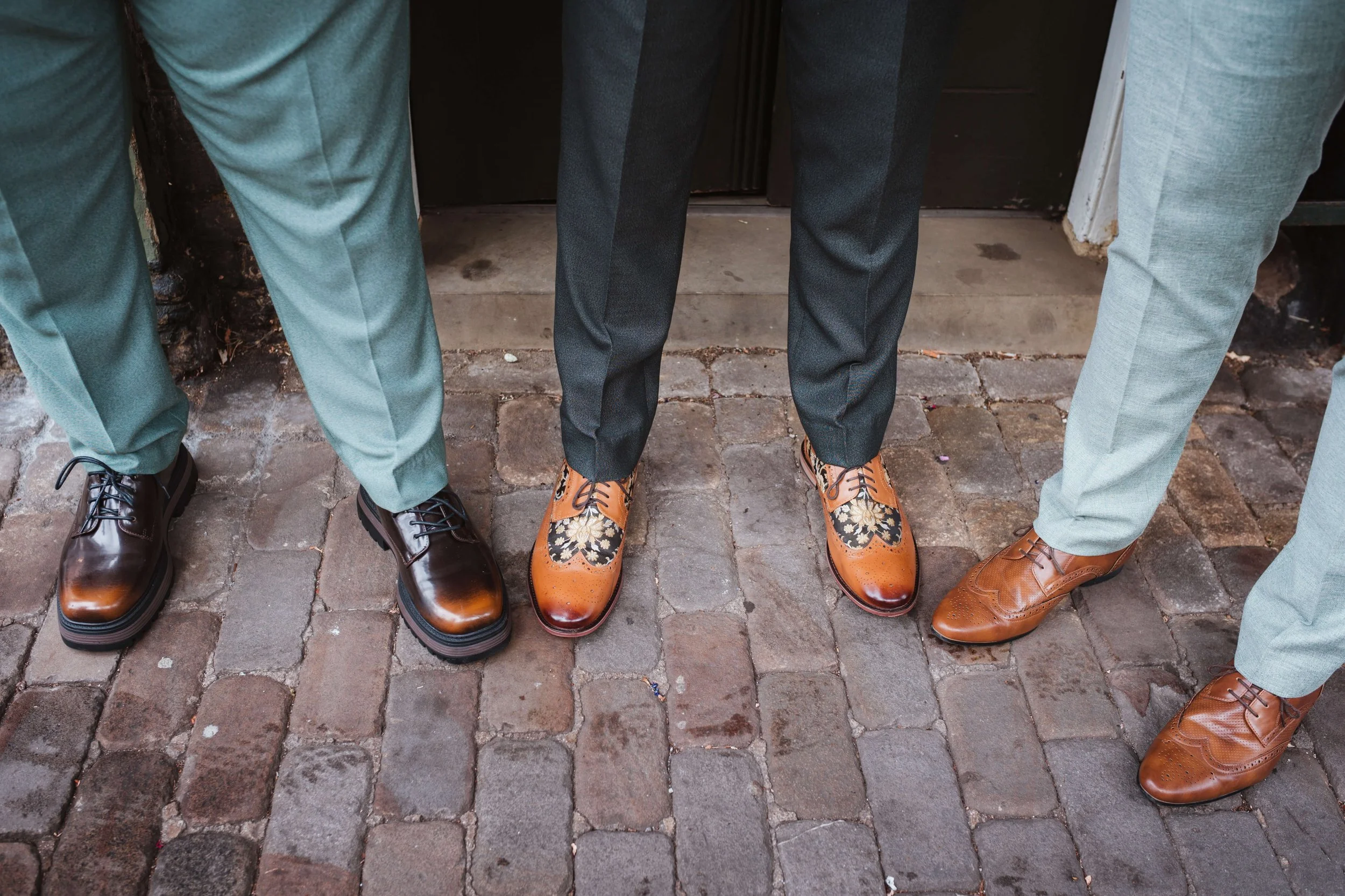 Close-up of three men's legs and shoes standing on a cobblestone sidewalk, with a black door in the background. The men are wearing dress shoes and suit pants in various shades of brown and gray.