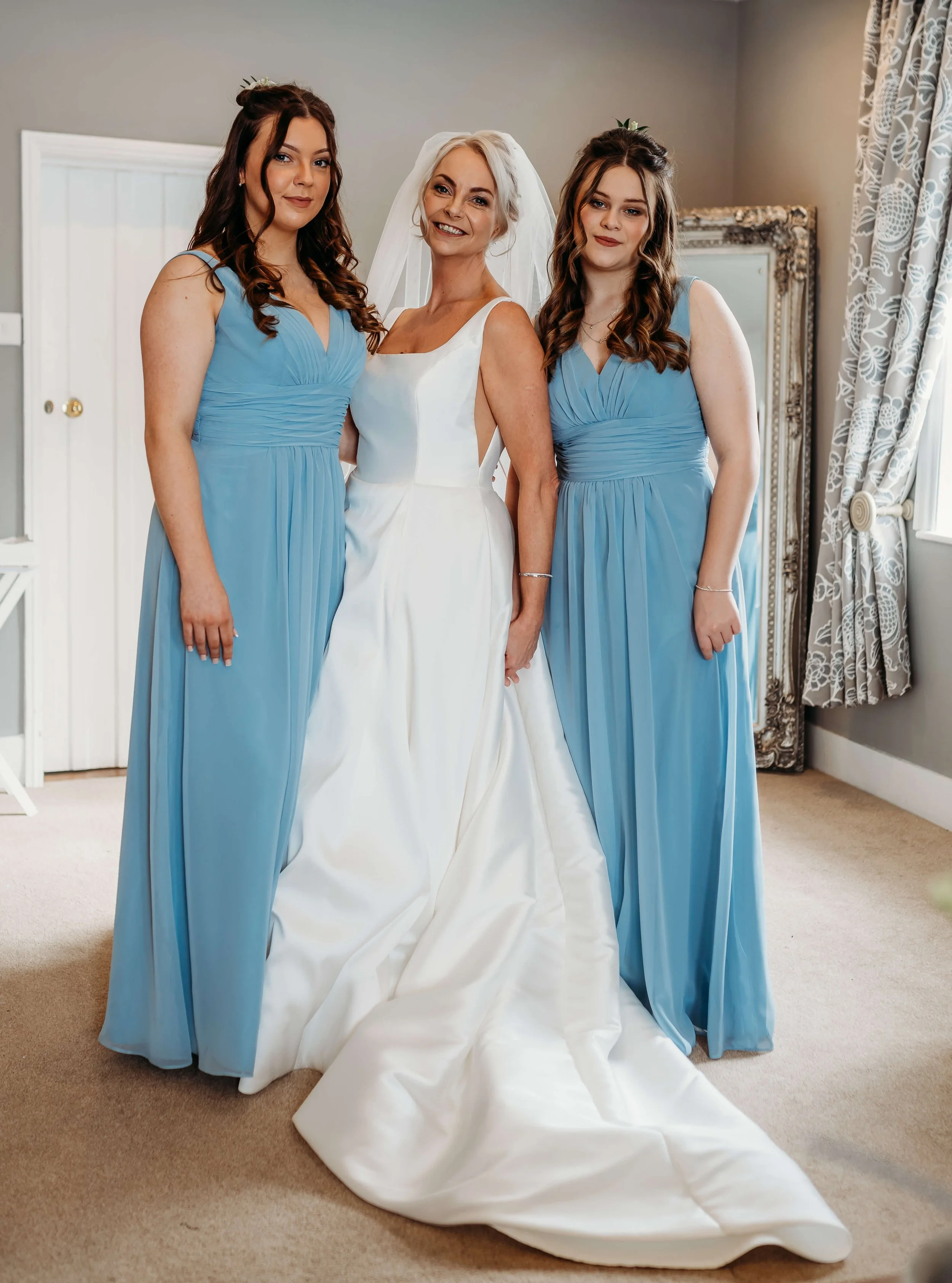 A bride in a white wedding dress standing between two bridesmaids in matching light blue dresses, all smiling inside a decorated room.