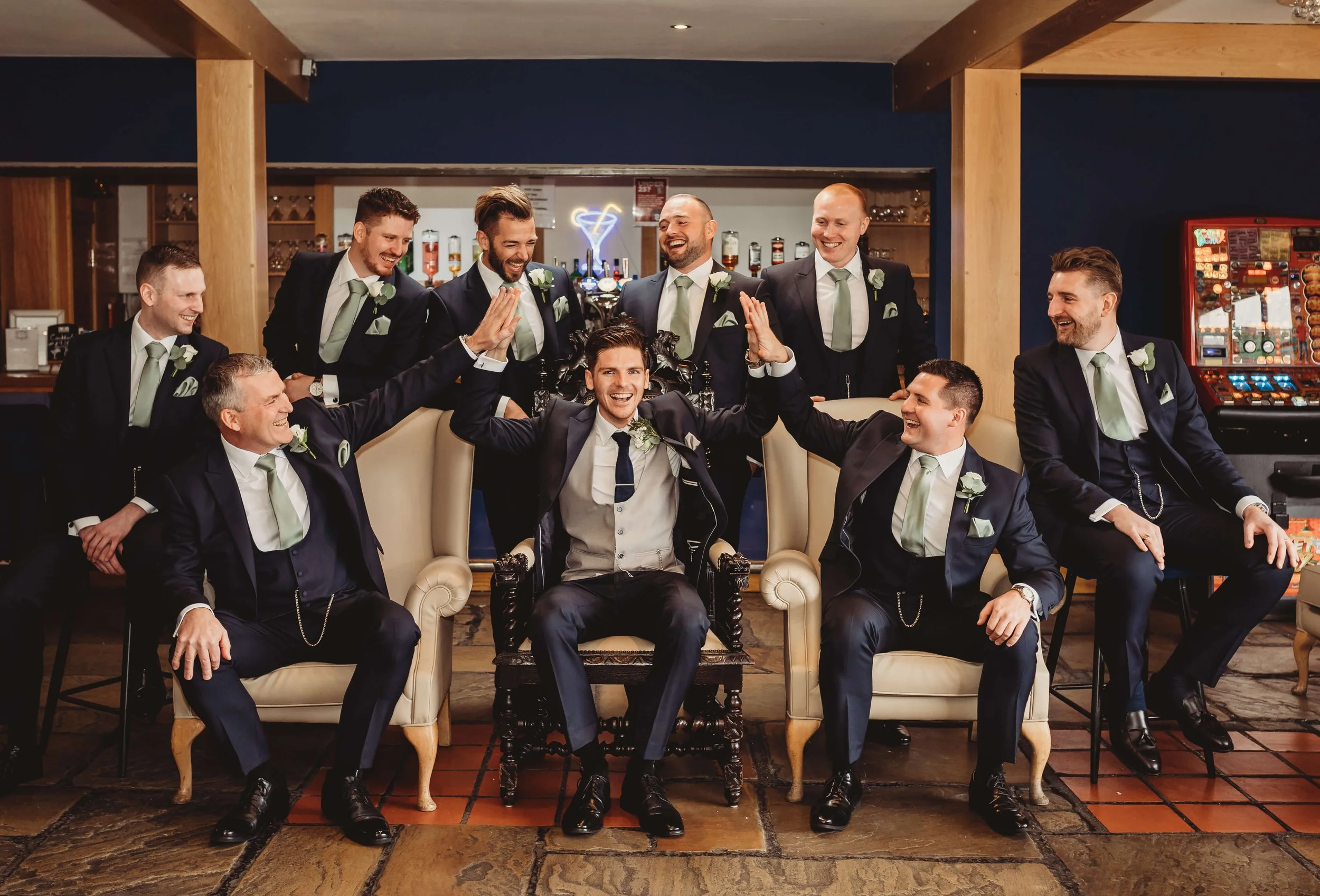 A group of ten groomsmen dressed in suits, some with light green ties and boutonnieres, celebrating with the groom seated in a central chair during a wedding or similar event.