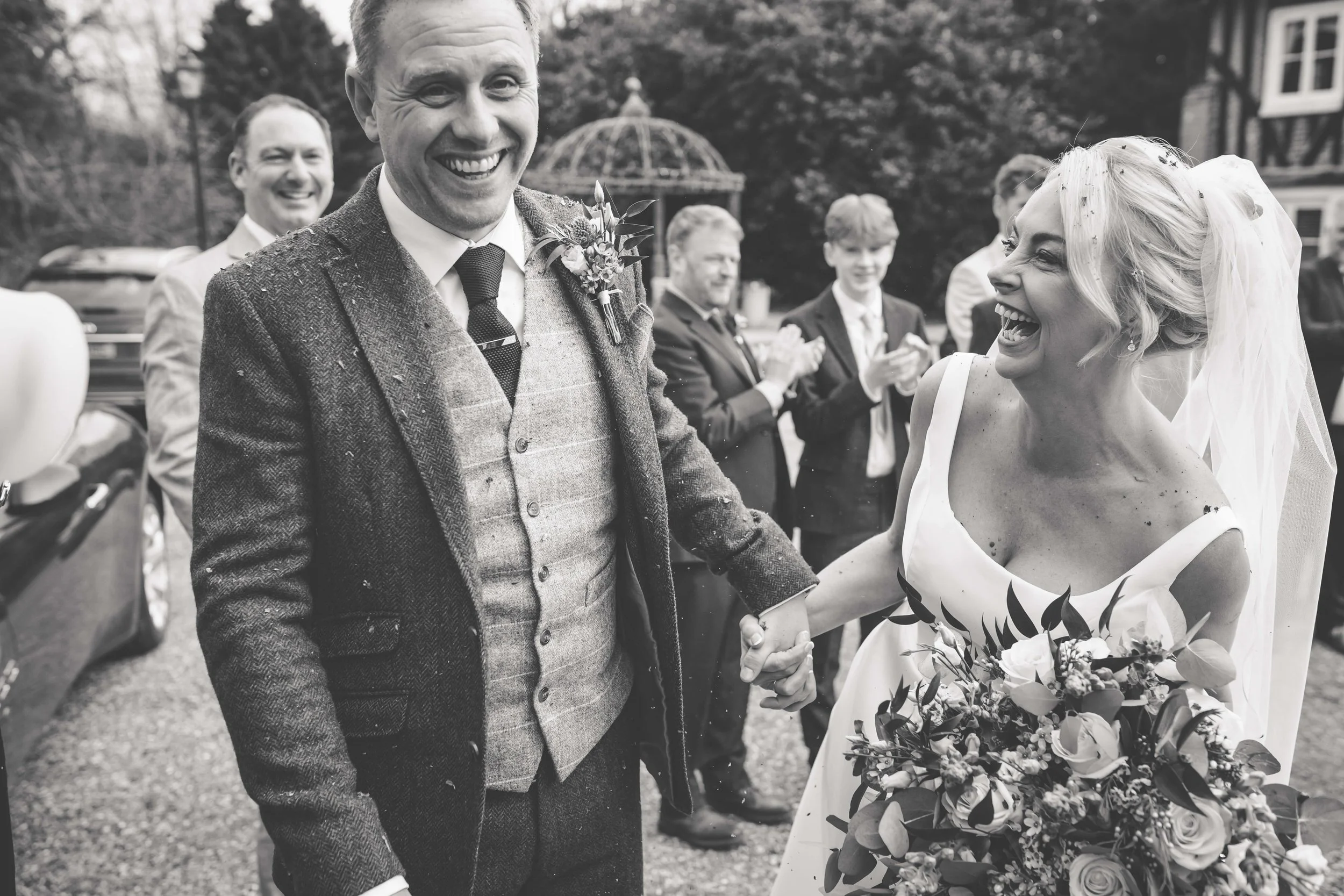 A bride and groom are holding hands and smiling during a wedding celebration outdoors, with a group of groomsmen or wedding guests clapping and smiling in the background.