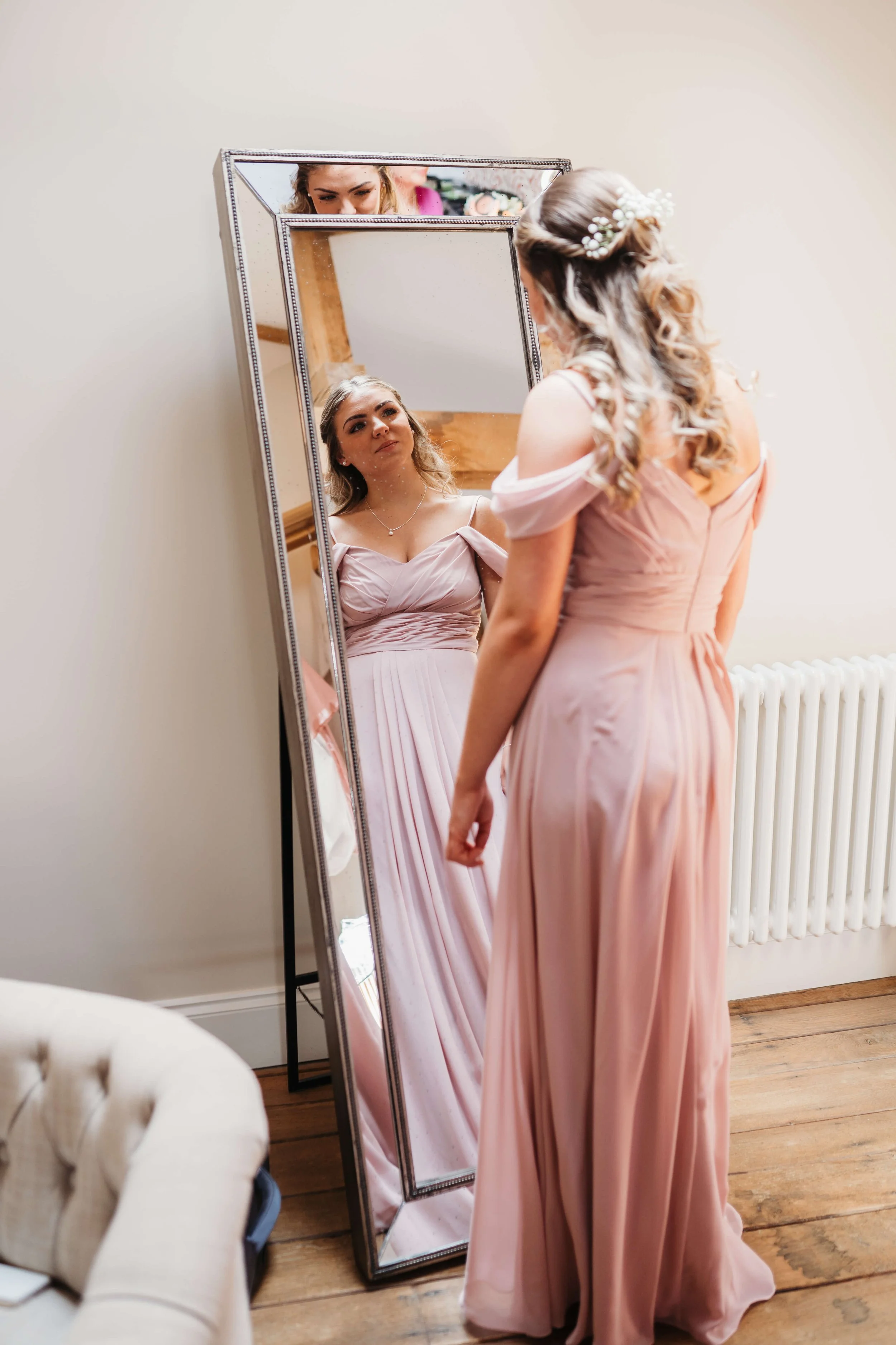 A woman in a pink dress with off-shoulder sleeves looks at herself in a full-length mirror, adjusting her dress. She has wavy hair styled with a floral headpiece. The room has wooden floors and a white wall with a radiator.
