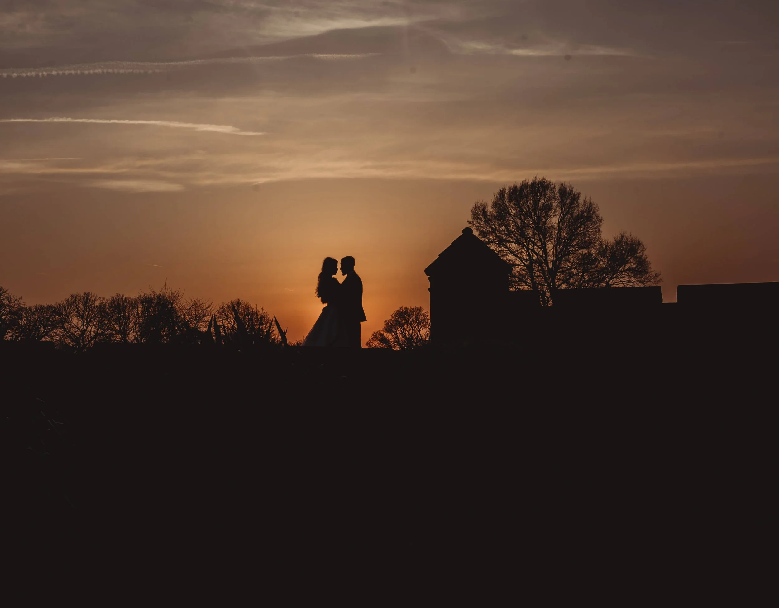 Silhouette of a couple embracing at sunset with trees and a small building in the background.