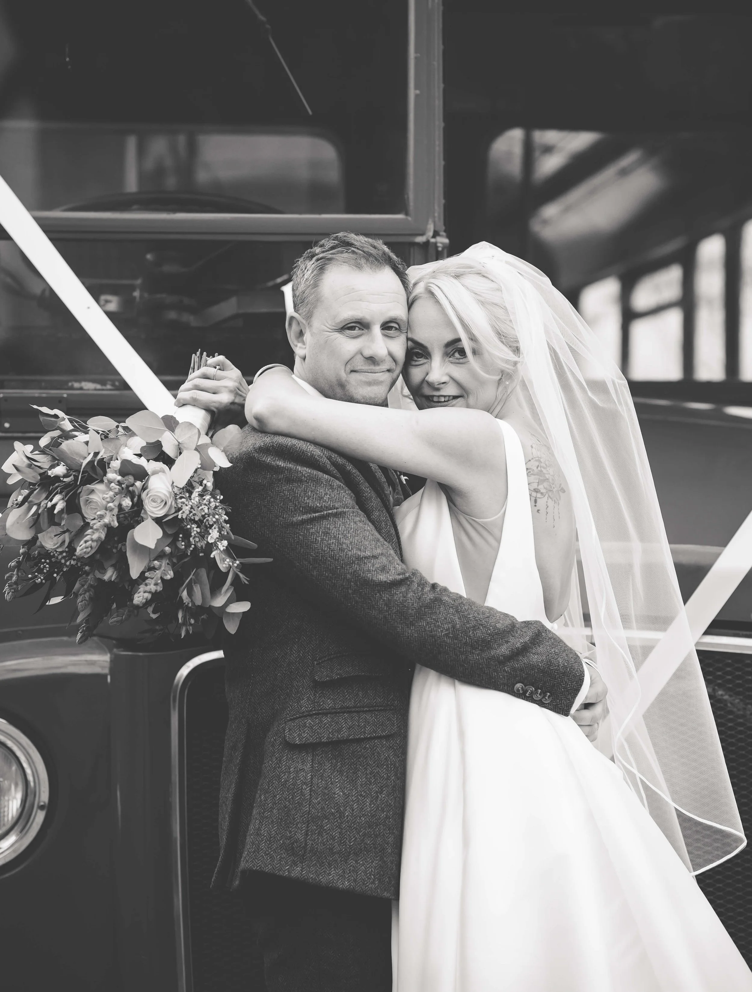 A black and white photo of a bride and groom embracing in front of a vehicle, with the bride holding a bouquet of flowers and smiling.