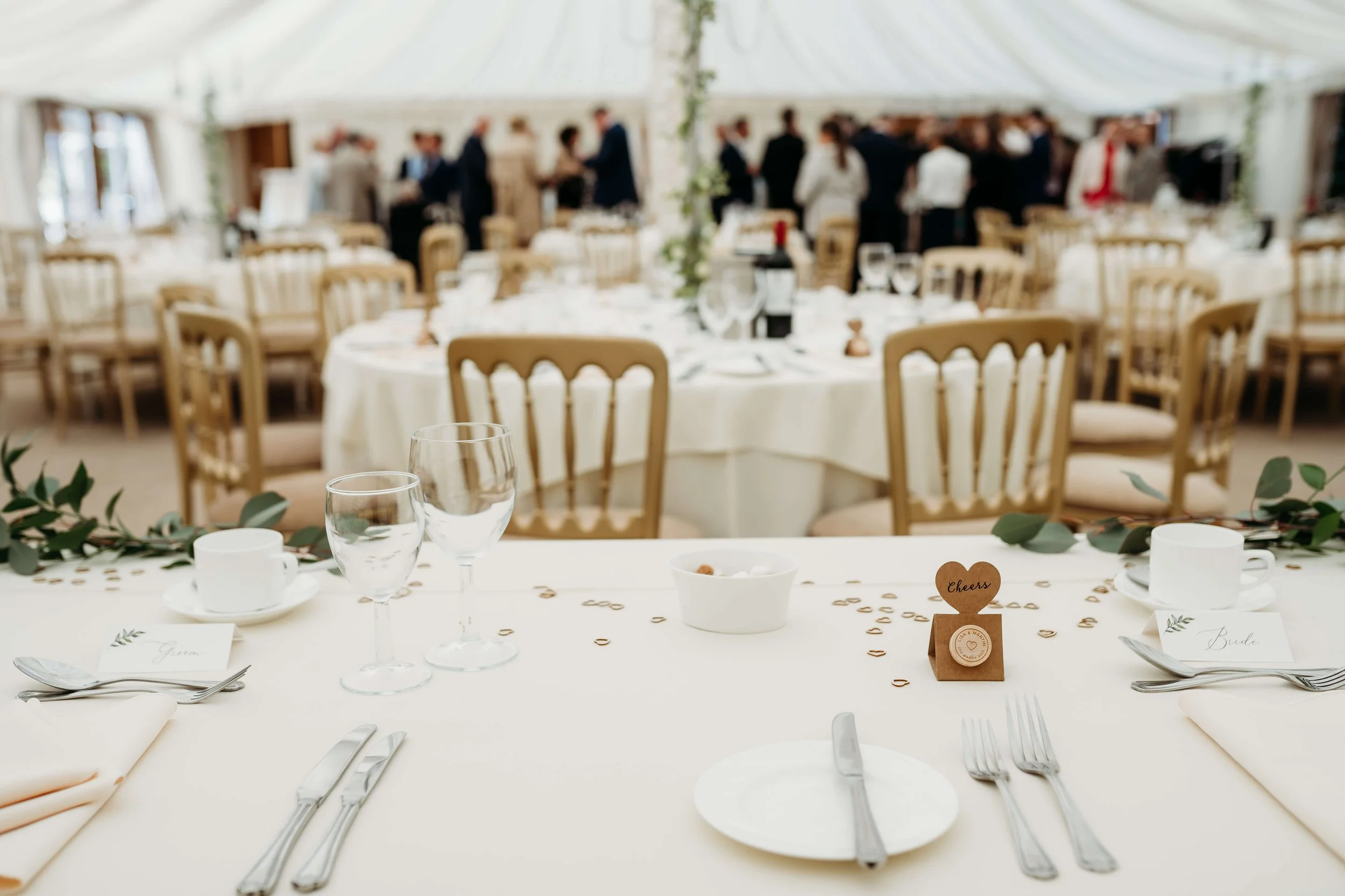 Wedding reception with decorated round tables, empty glassware, cutlery, and signs for the bride and groom, set in a well-lit tented venue with guests in the background.