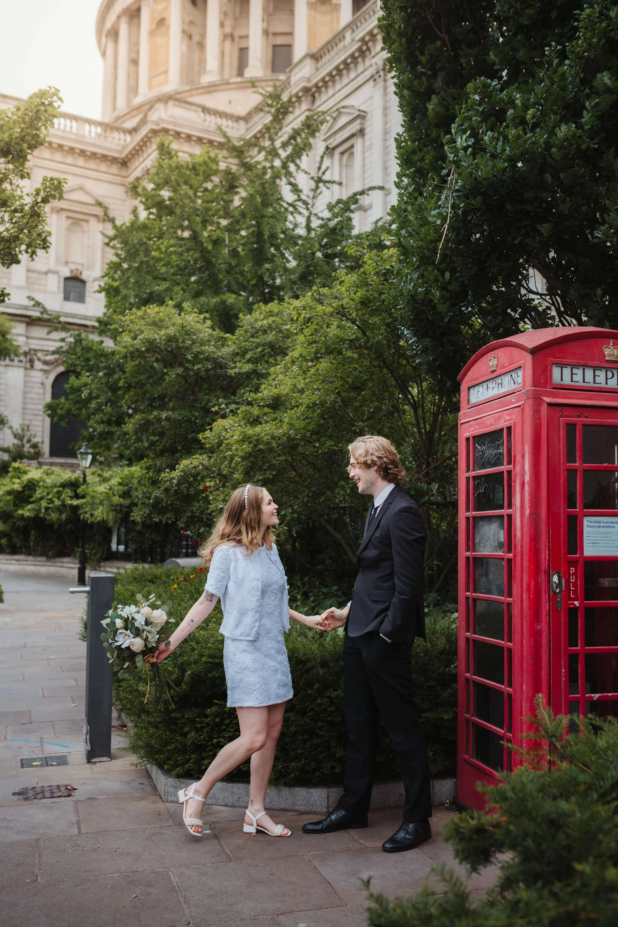 A couple dressed in formal attire holding hands and smiling outside near a red telephone booth, with a historic building and green trees in the background.