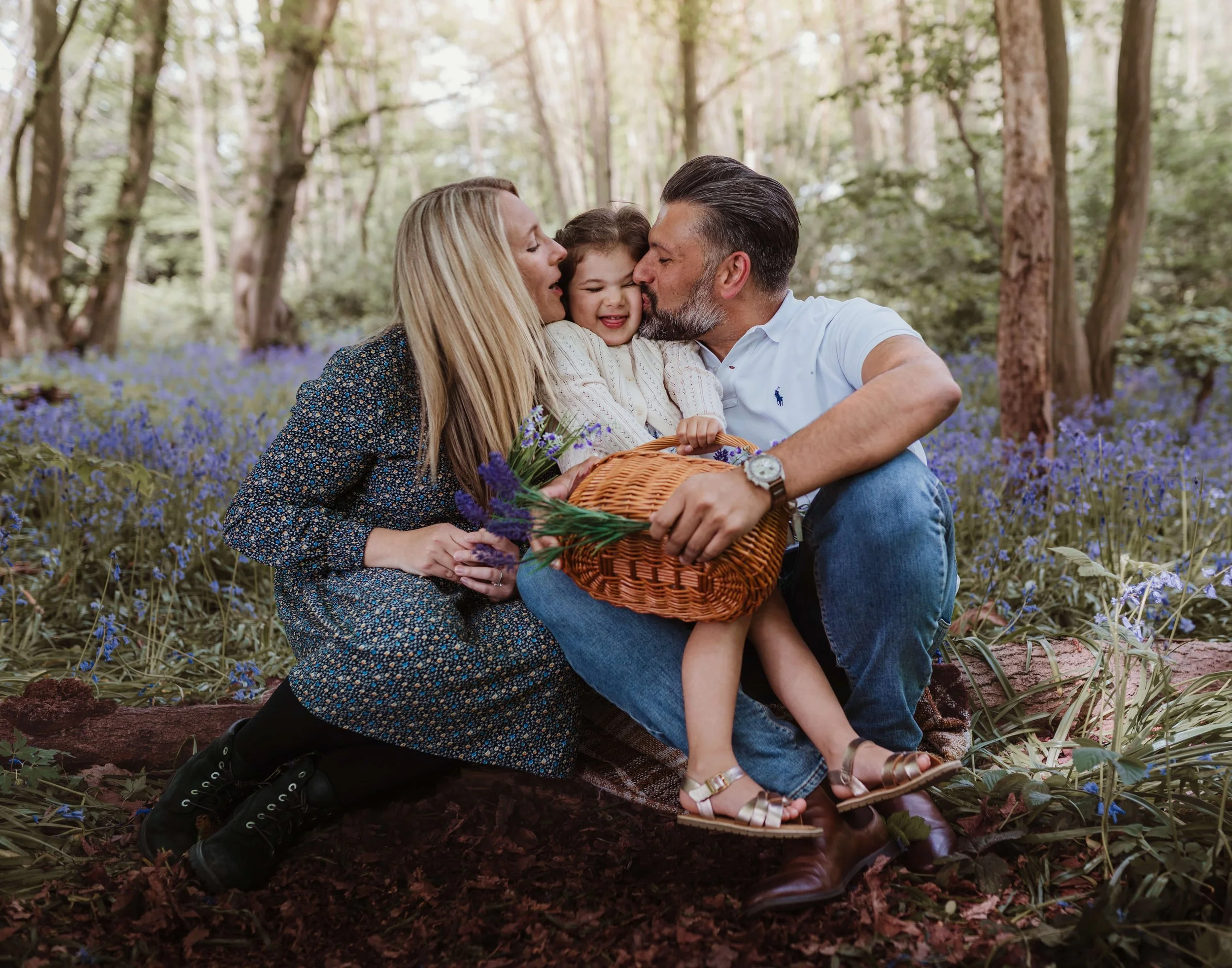 A family of three, a man, woman, and a young girl, sitting on a blanket in a forest with purple flowers, sharing a kiss and smiling, with the woman holding a basket of flowers.