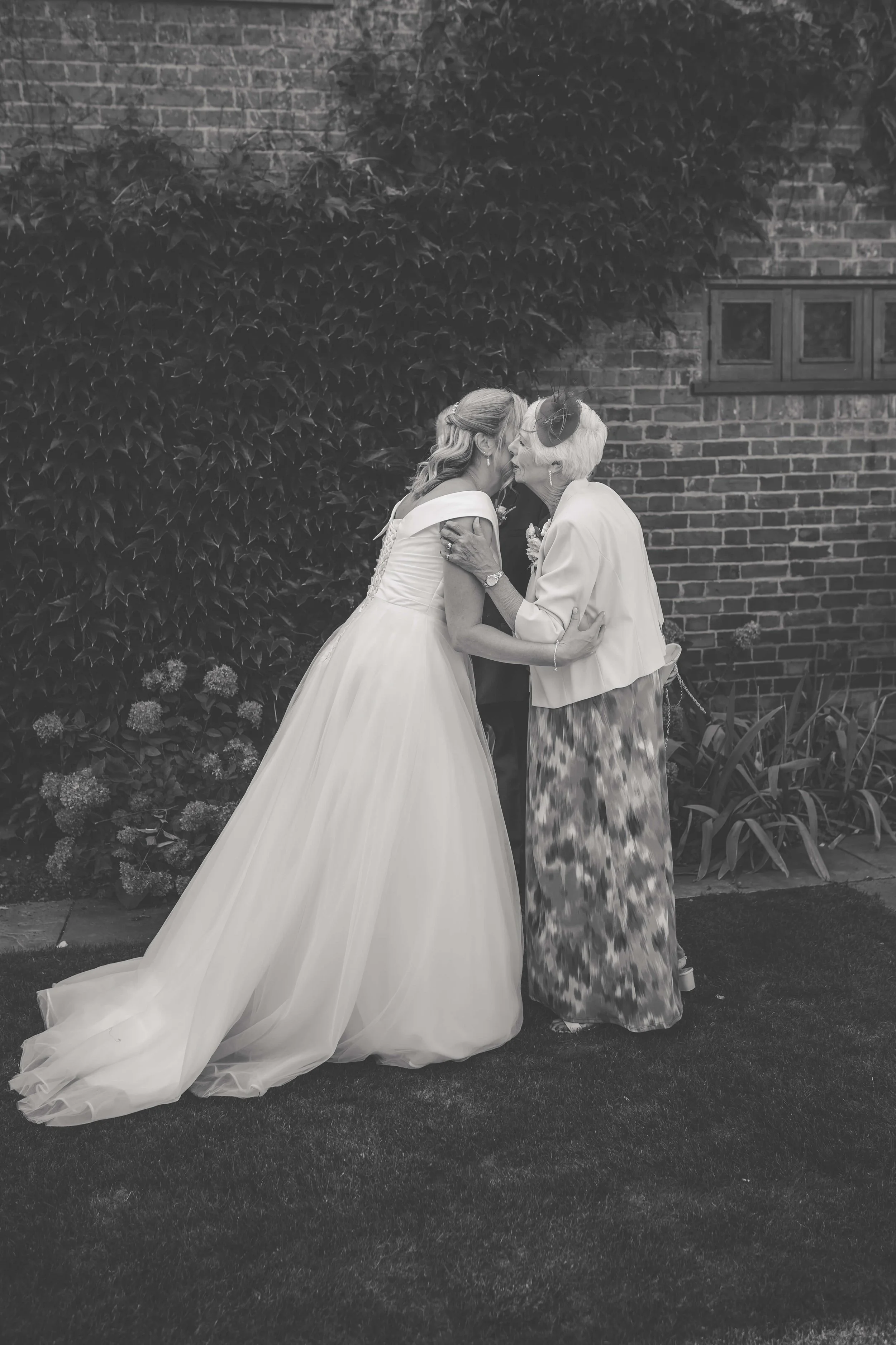 A wedding bride in a white gown sharing a kiss with an elderly woman, possibly her grandmother, outdoors beside a brick wall and greenery.