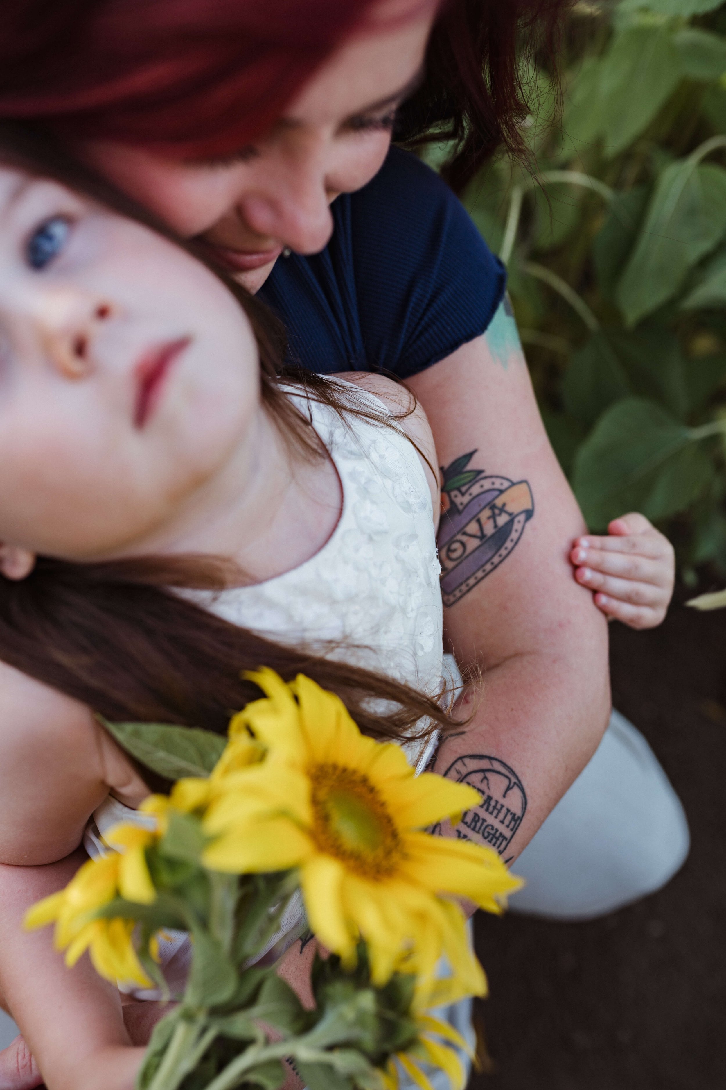 A woman with red hair holding a young girl with brown hair, both surrounded by greenery. The girl holds a bouquet of yellow sunflowers.