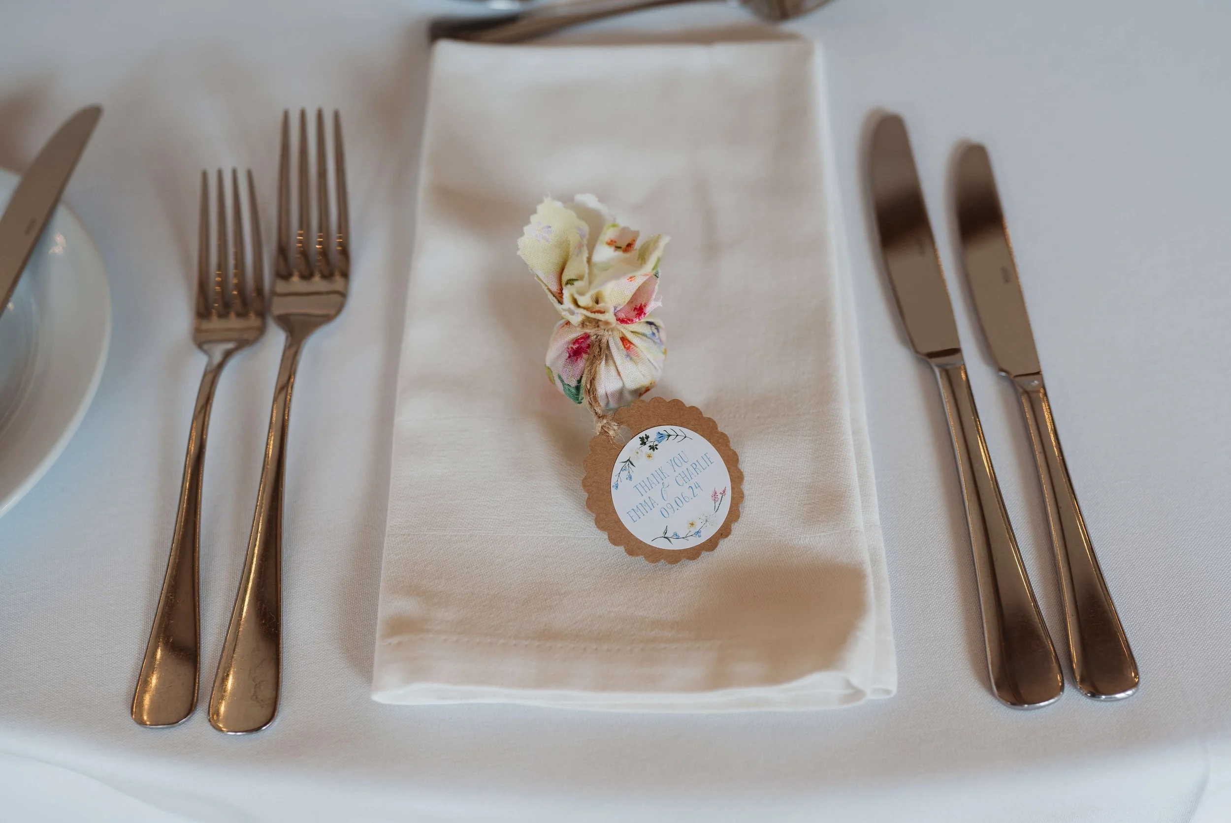 Elegant table setting with a folded white napkin, two forks on the left, two knives on the right, and a floral fabric-wrapped favor with a thank you tag in the center.