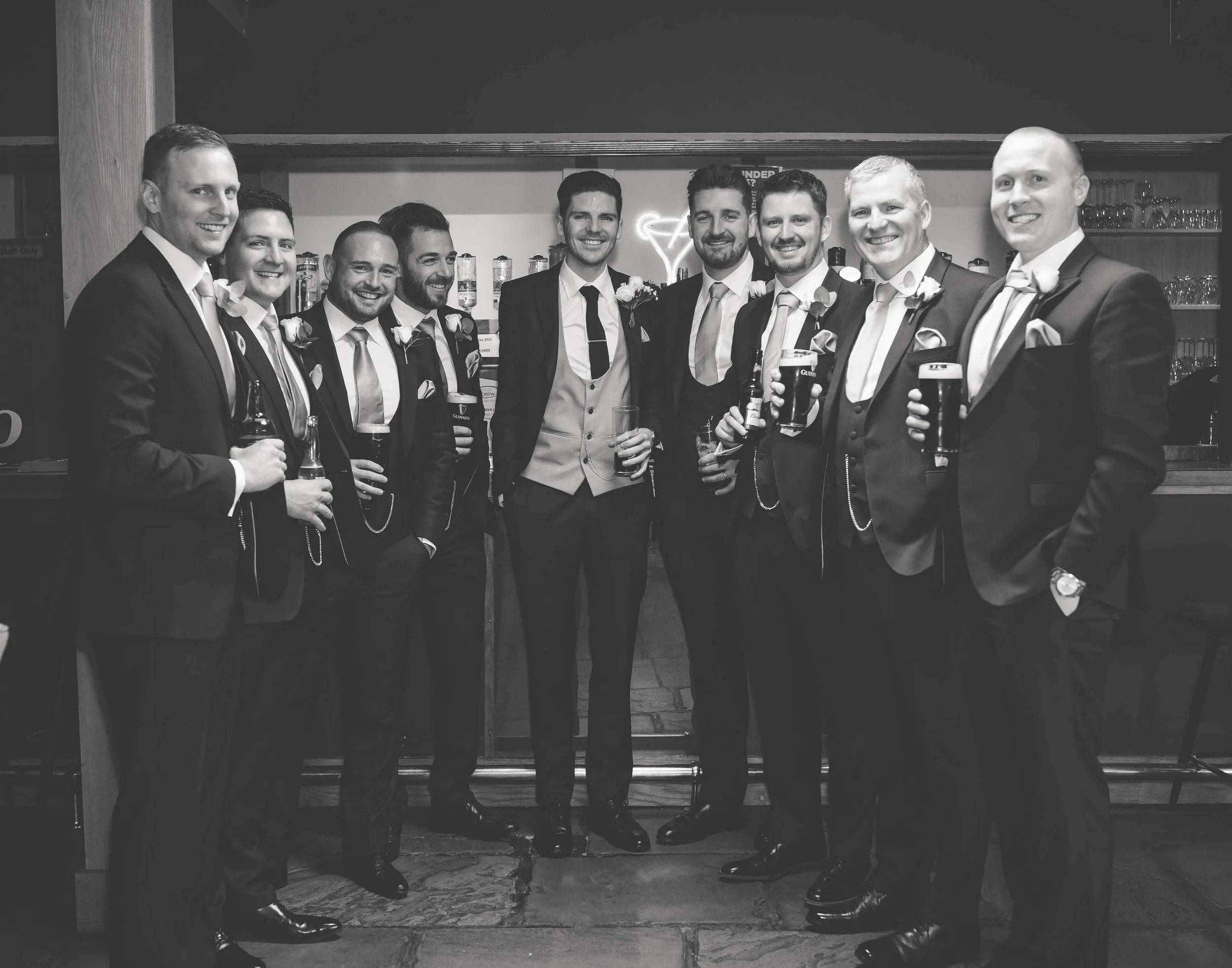 A group of eleven men in suits at a social gathering, possibly a wedding, holding drinks and smiling.