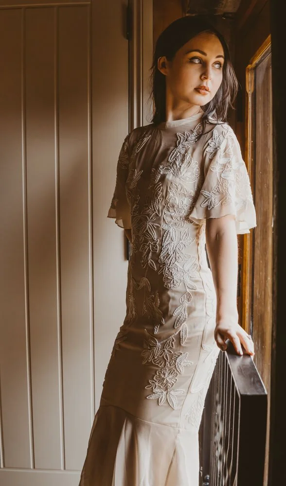 A woman wearing a beige dress with floral embroidery, standing by a window and looking outside.
