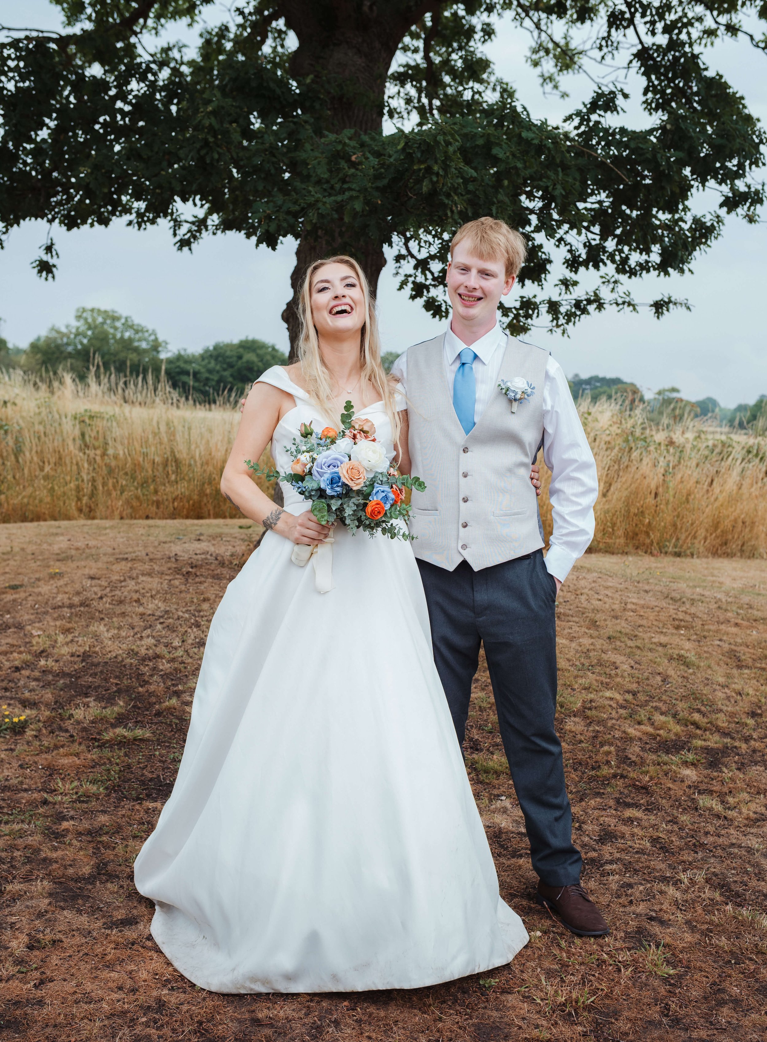 A wedding couple standing outdoors under a large tree, with tall grass in the background. The bride is wearing a white wedding gown and holding a bouquet of flowers, and the groom is dressed in a light vest, white shirt, and blue tie. They are smilin