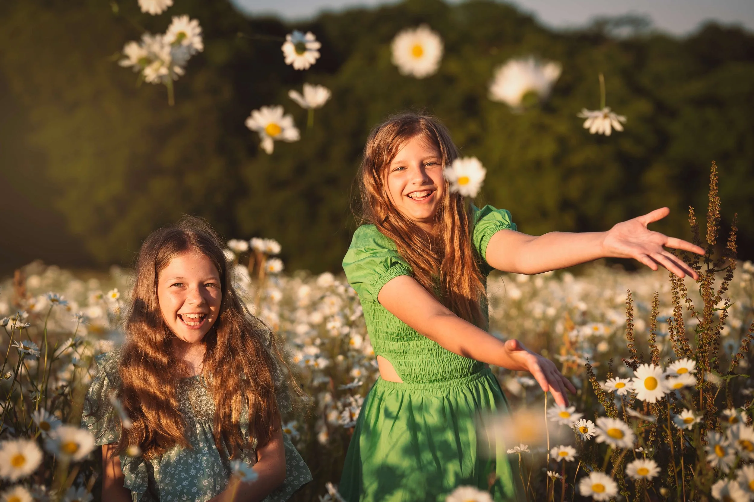 Two young girls with long wavy hair playing in a field of daisies during sunset, one wearing a green dress and smiling, the other wearing a gray dress and laughing.