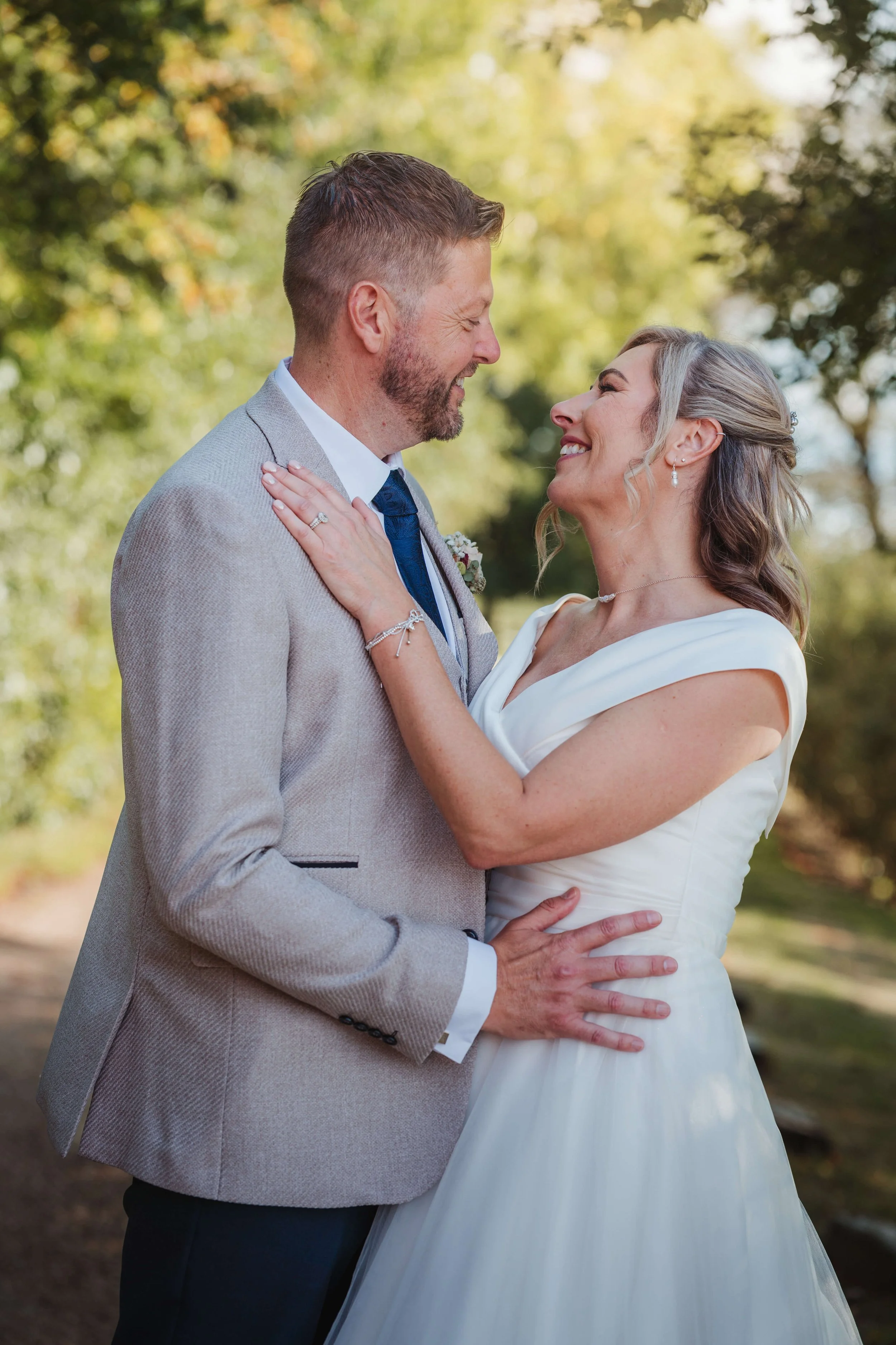 A smiling bride and groom looking into each other's eyes outdoors, with trees and sunlight in the background.