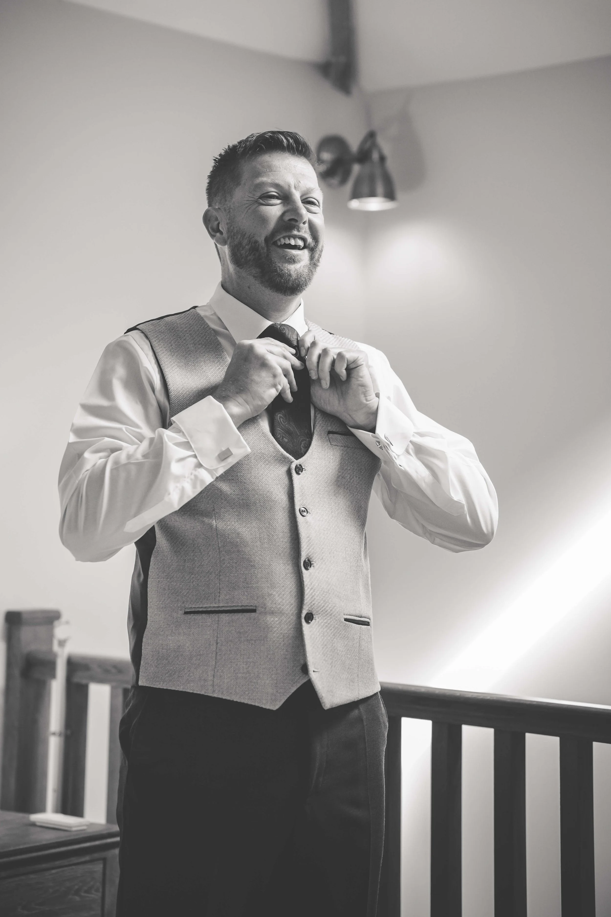 A man with a beard smiling and adjusting his tie, dressed in formal attire with a vest, in an indoor setting.