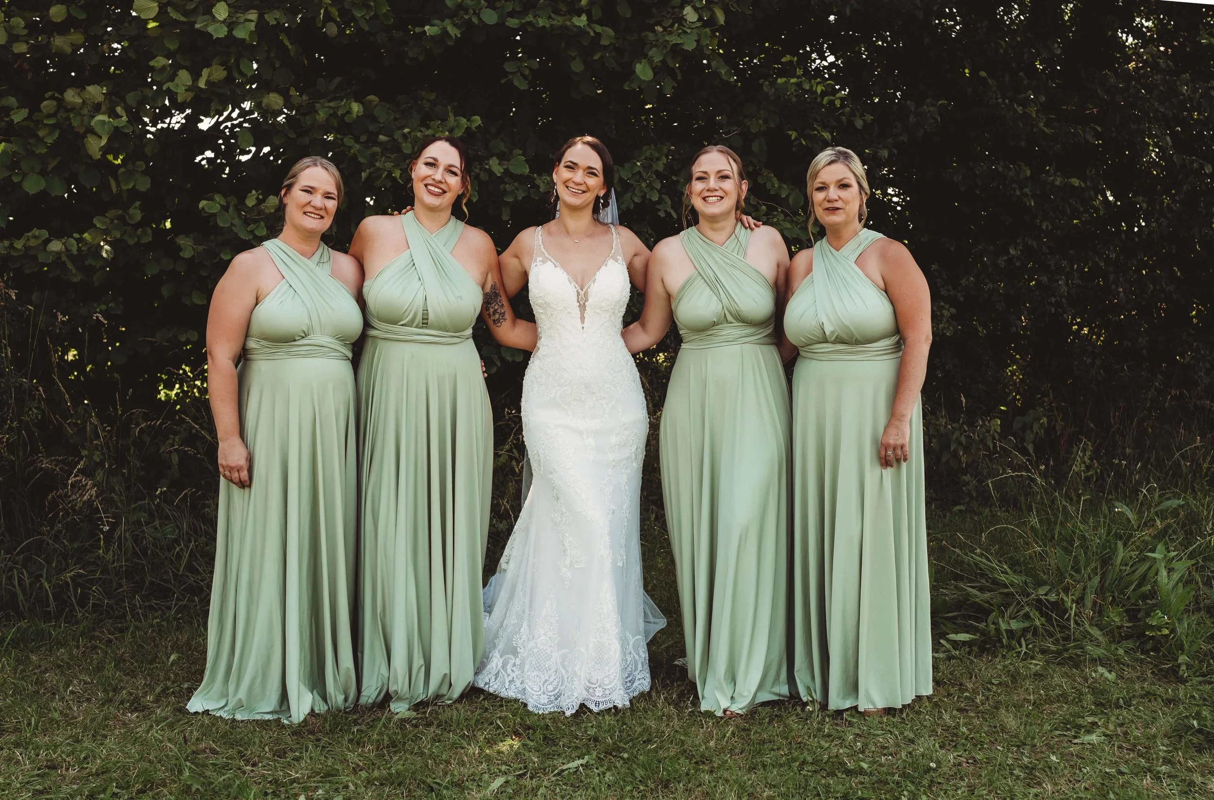 A bride in a white lace wedding gown standing with five bridesmaids in matching sage green dresses outdoors, smiling in front of a leafy background.