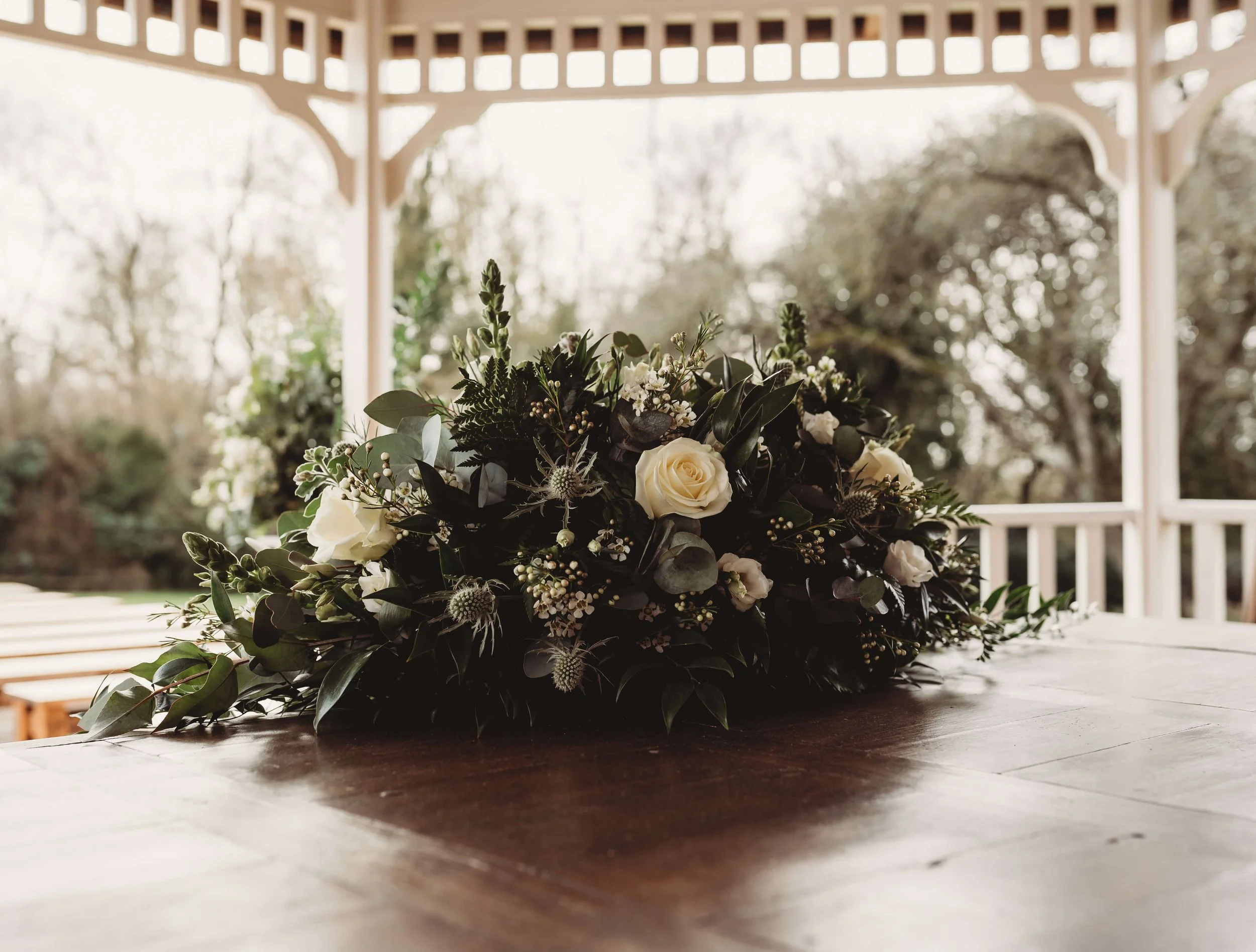 A bouquet of white roses and greenery on a wooden table with a white gazebo and trees in the background.