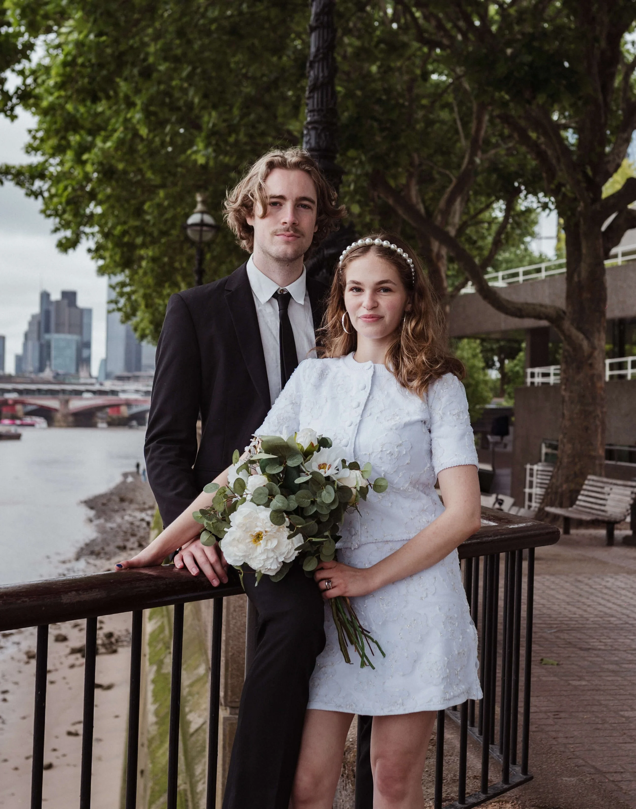 A young couple dressed in wedding attire posing by a river, with city buildings in the background, trees, and a promenade.