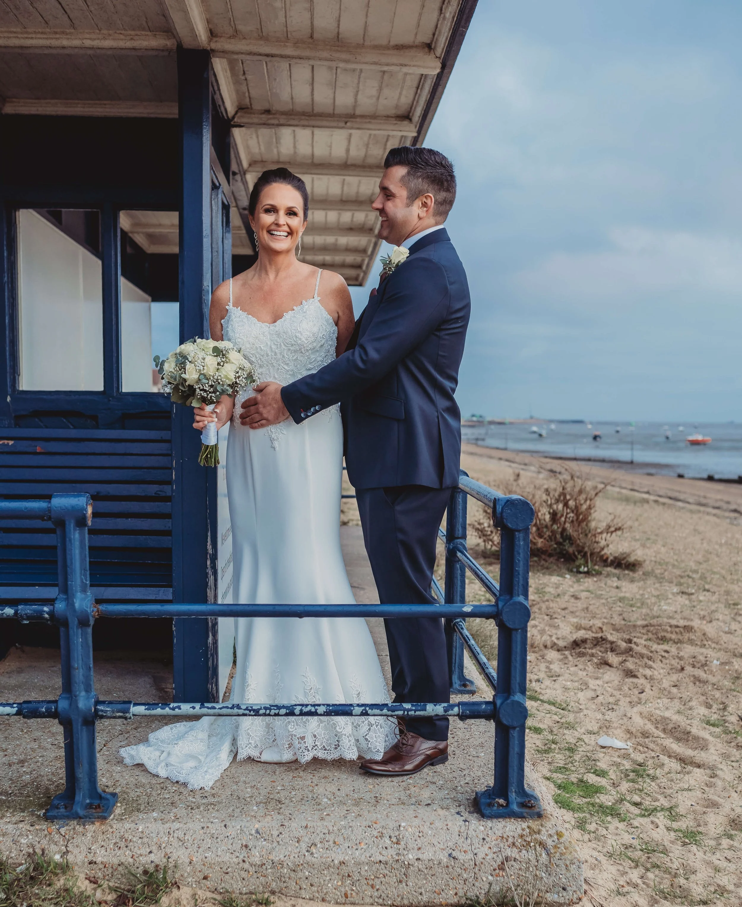 A bride and groom standing on a beachside walkway, smiling at each other. The bride holds a bouquet of white flowers, and they are positioned near a blue beach hut with boats and the ocean in the background.